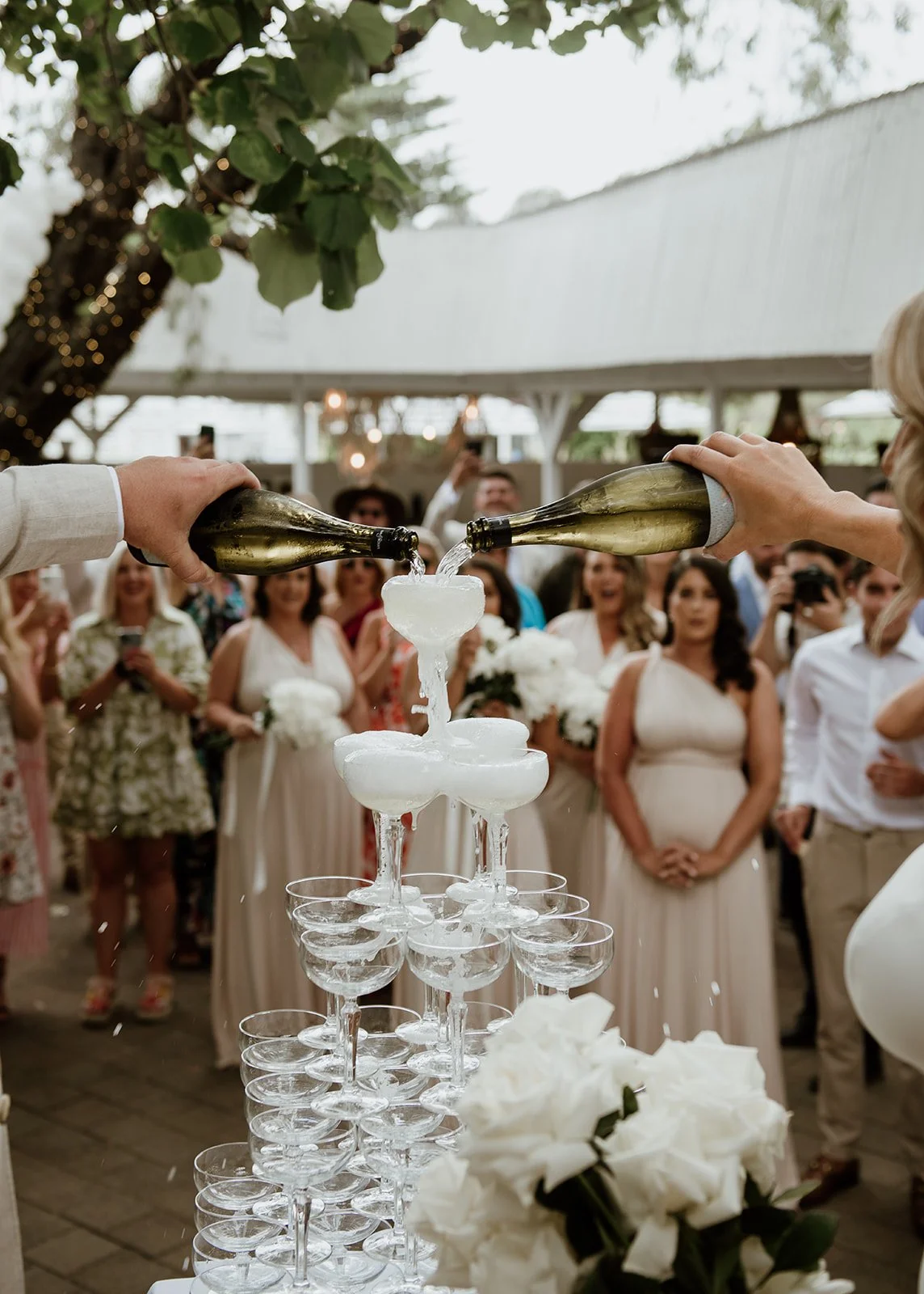 A wedding celebration where two people are pouring champagne into a pyramid of glass champagne coupes, with guests watching and taking photos in the background.