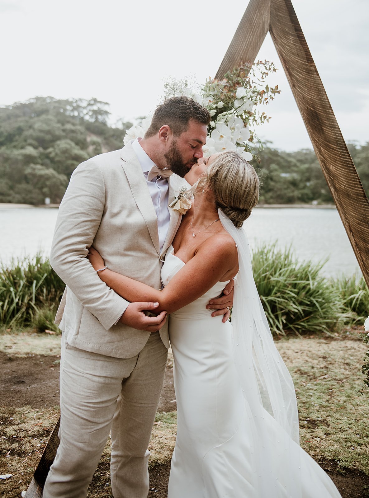 A wedding couple, a man and a woman, sharing a kiss outdoors near a lake with trees in the background. They stand in front of a wooden triangular arch decorated with white flowers and greenery.