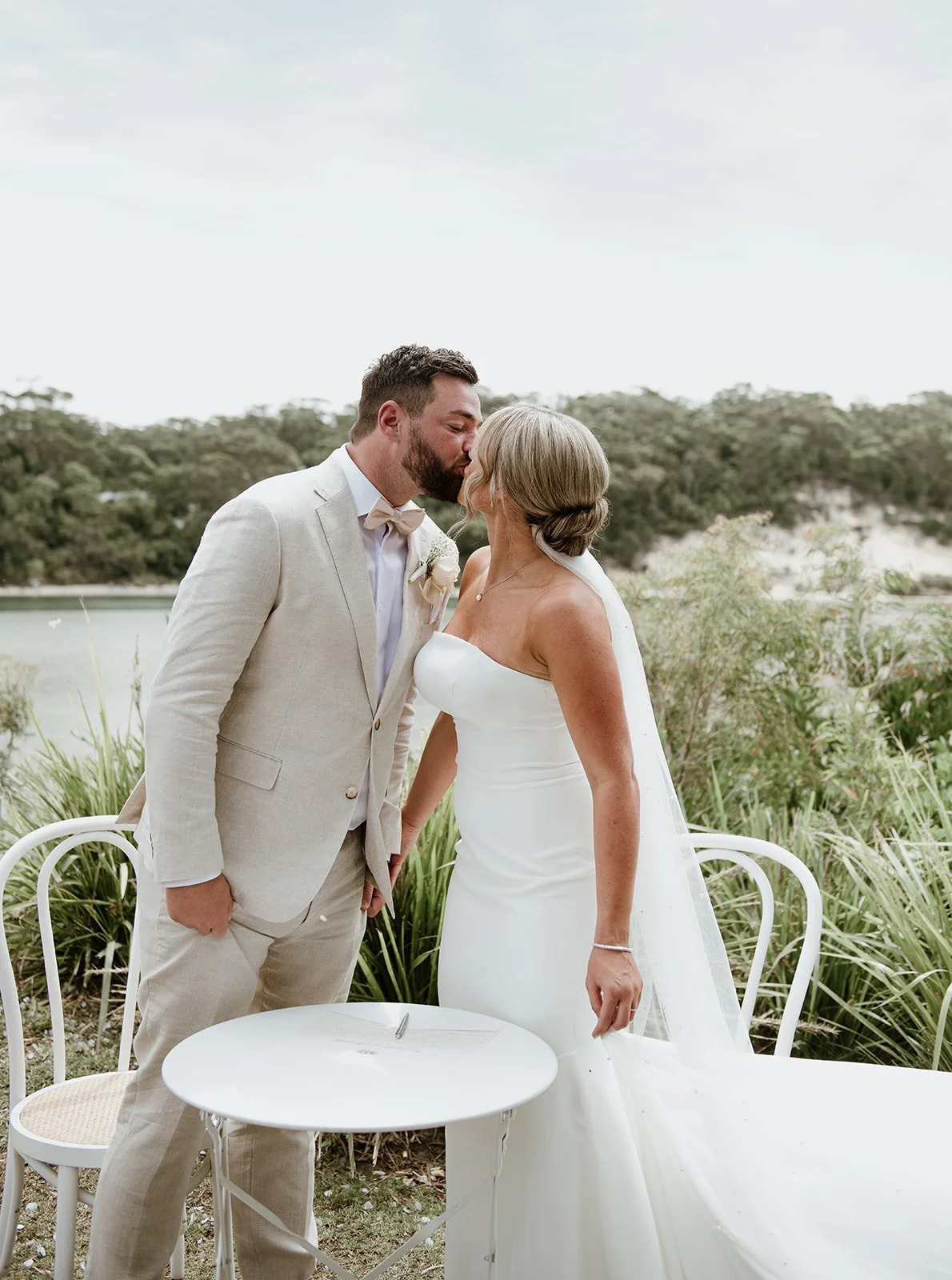 A bride and groom sharing a kiss outdoors during their wedding ceremony, with greenery and a body of water in the background.