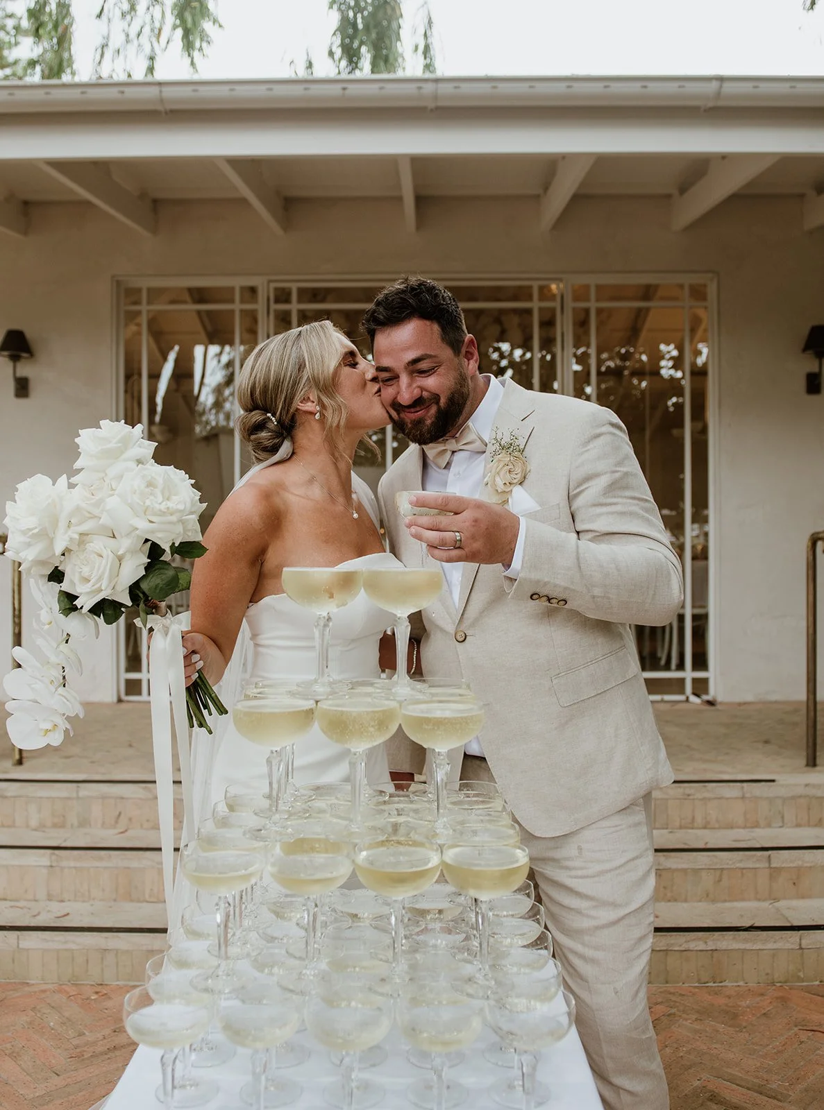 A bride and groom celebrating at their wedding reception, standing behind a tower of champagne glasses, with the bride holding a bouquet of white flowers and kissing the groom on the cheek.