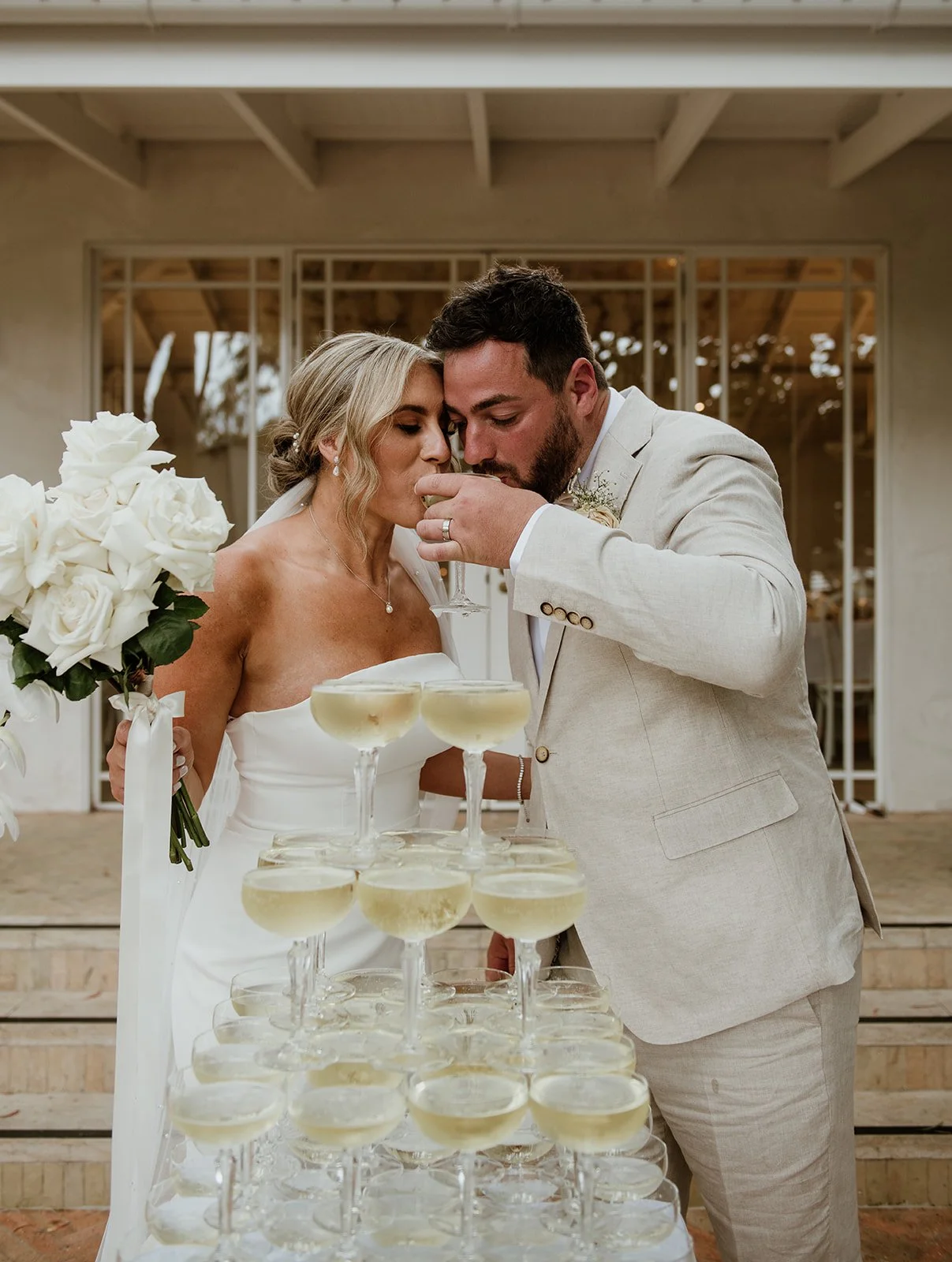 A bride and groom sharing a toast at their wedding reception, pouring champagne into glasses, with a cascade of champagne glasses set up in front of them