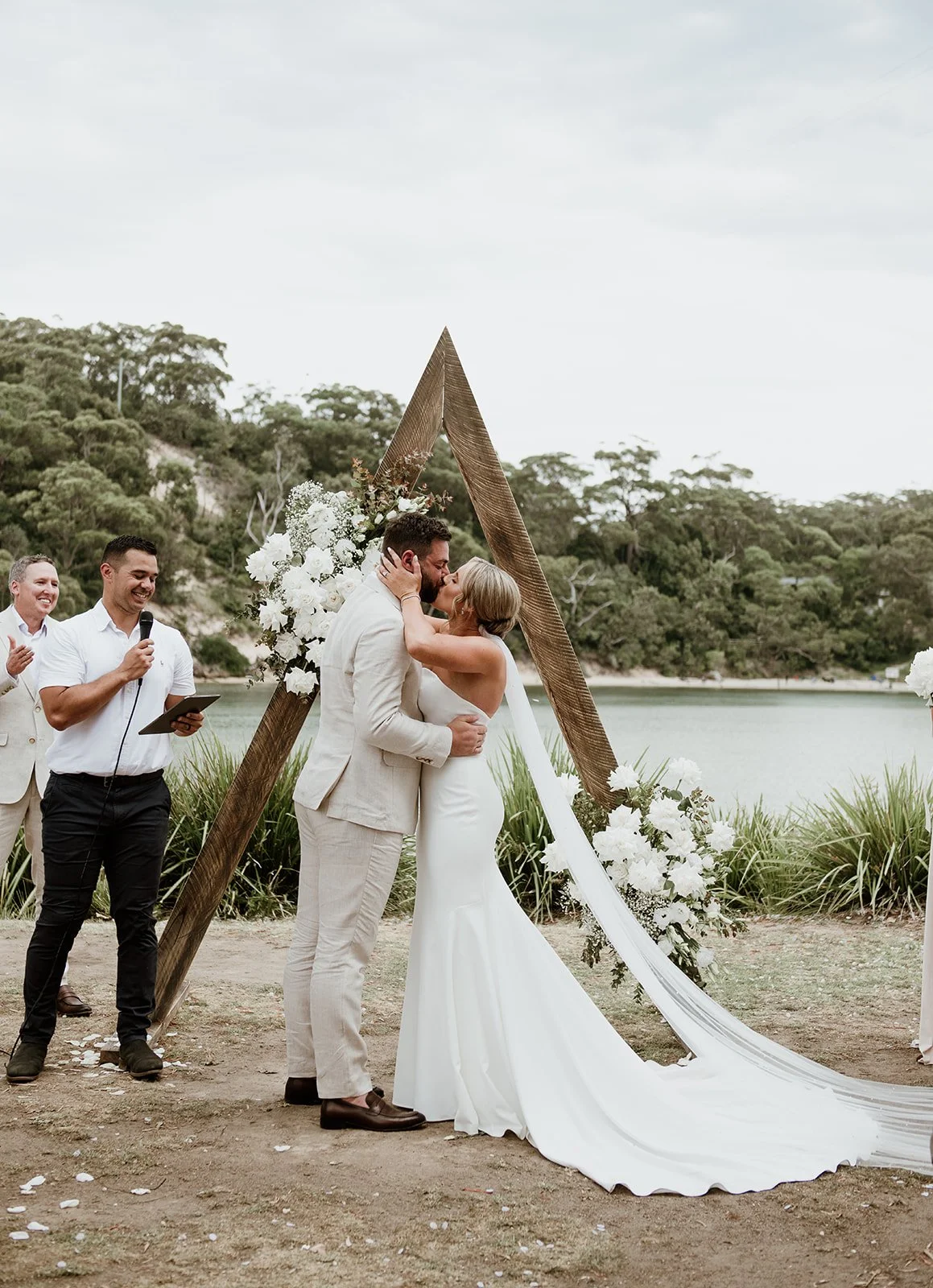 A couple in wedding attire sharing a kiss during their outdoor wedding ceremony near water, with officiants and a decorative wooden arch with white flowers and greenery in the background.