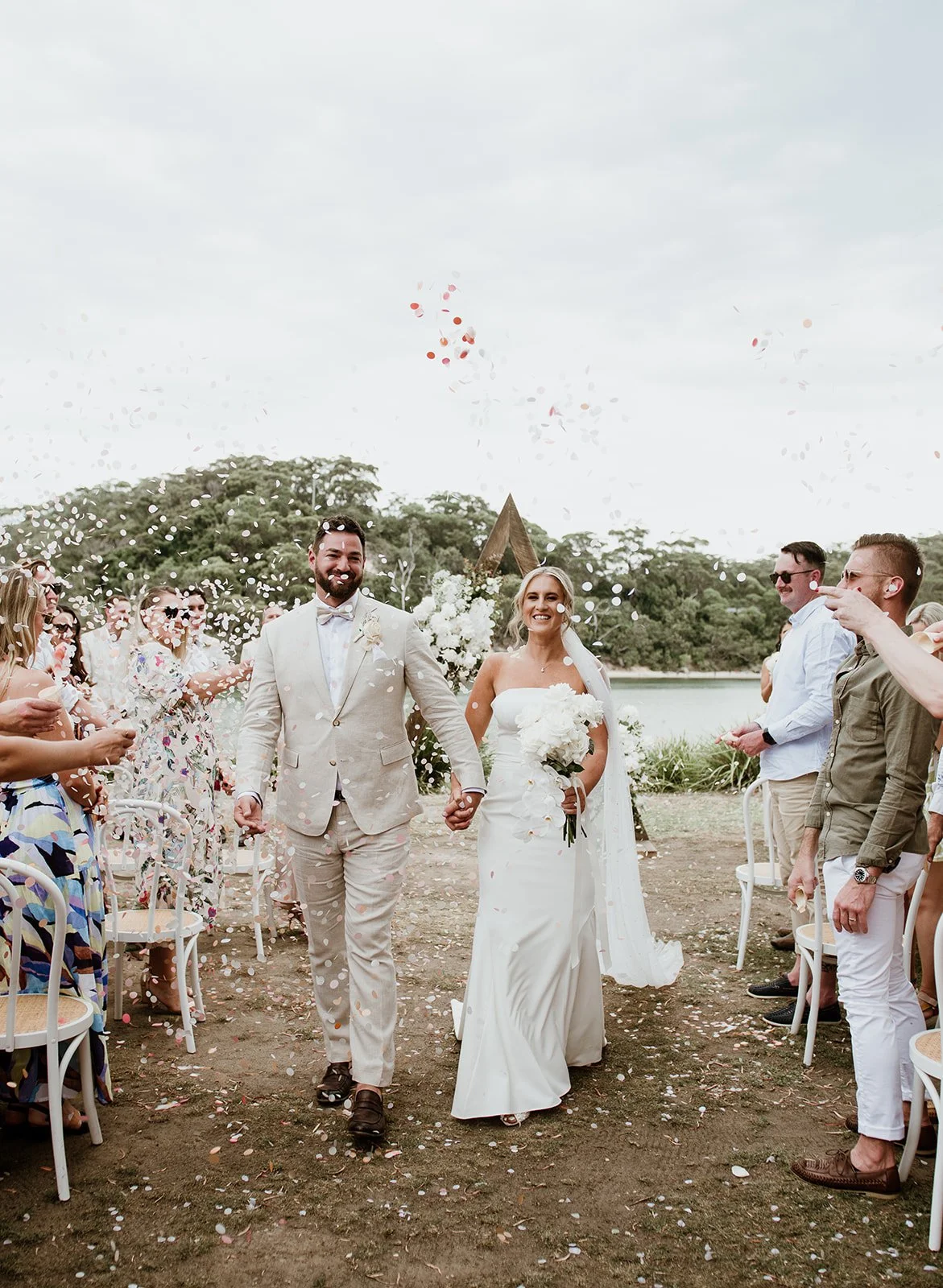A bride and groom walking hand in hand down the outdoor aisle after their wedding ceremony, surrounded by friends and family, with balloons and confetti in the air near a lake and trees in the background.