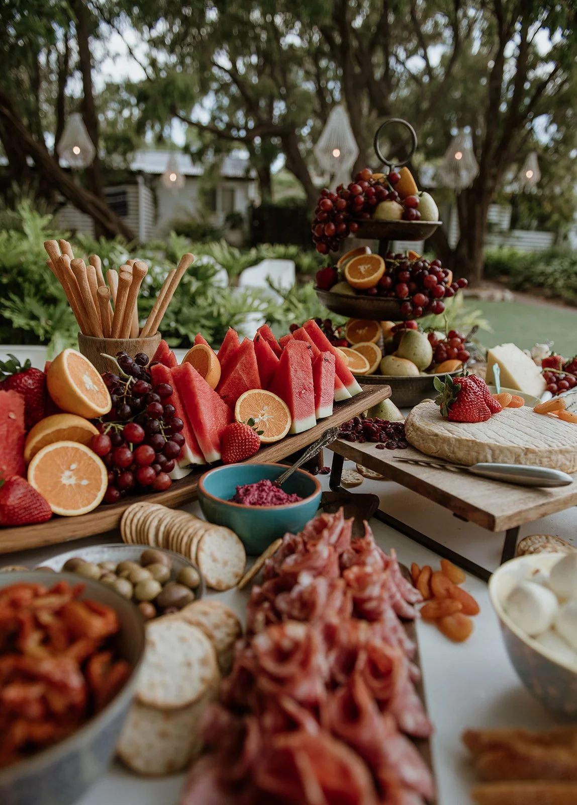A colorful outdoor wedding fruit and snack table with watermelon slices, citrus halves, grapes, strawberries, cheese, crackers, and dips, with a backdrop of trees and hanging lights.