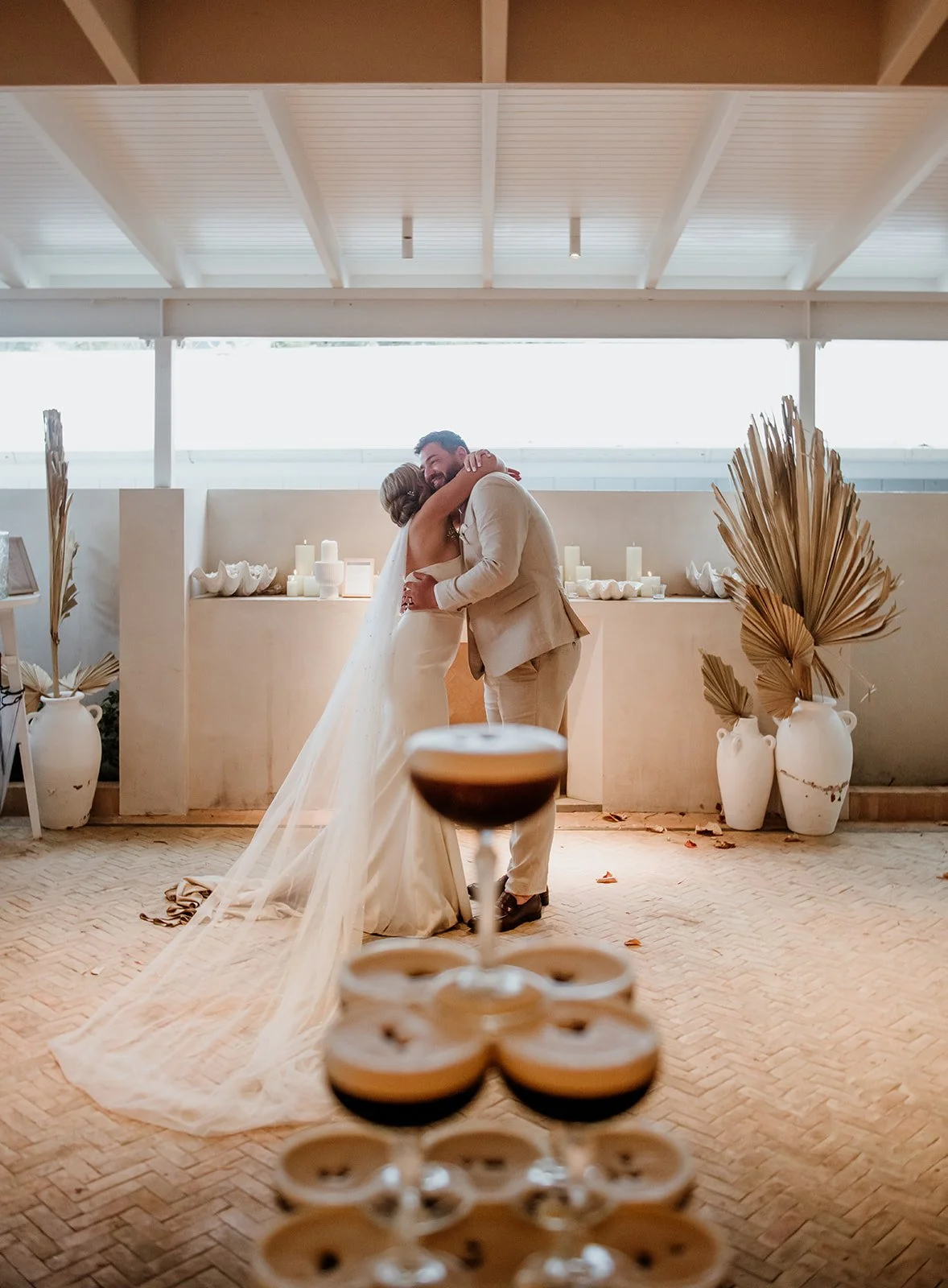 A bride and groom hugging during their wedding reception, with a tower of drinks in the foreground and decorative vases with dried leaves in the background.