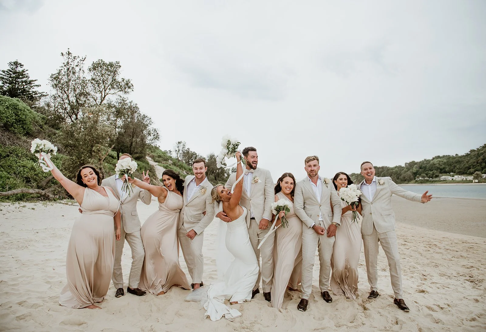 A wedding party of nine people standing on a beach with ocean and trees in the background, smiling and celebrating, the bride and groom in the center with bridesmaids and groomsmen around them holding bouquets.
