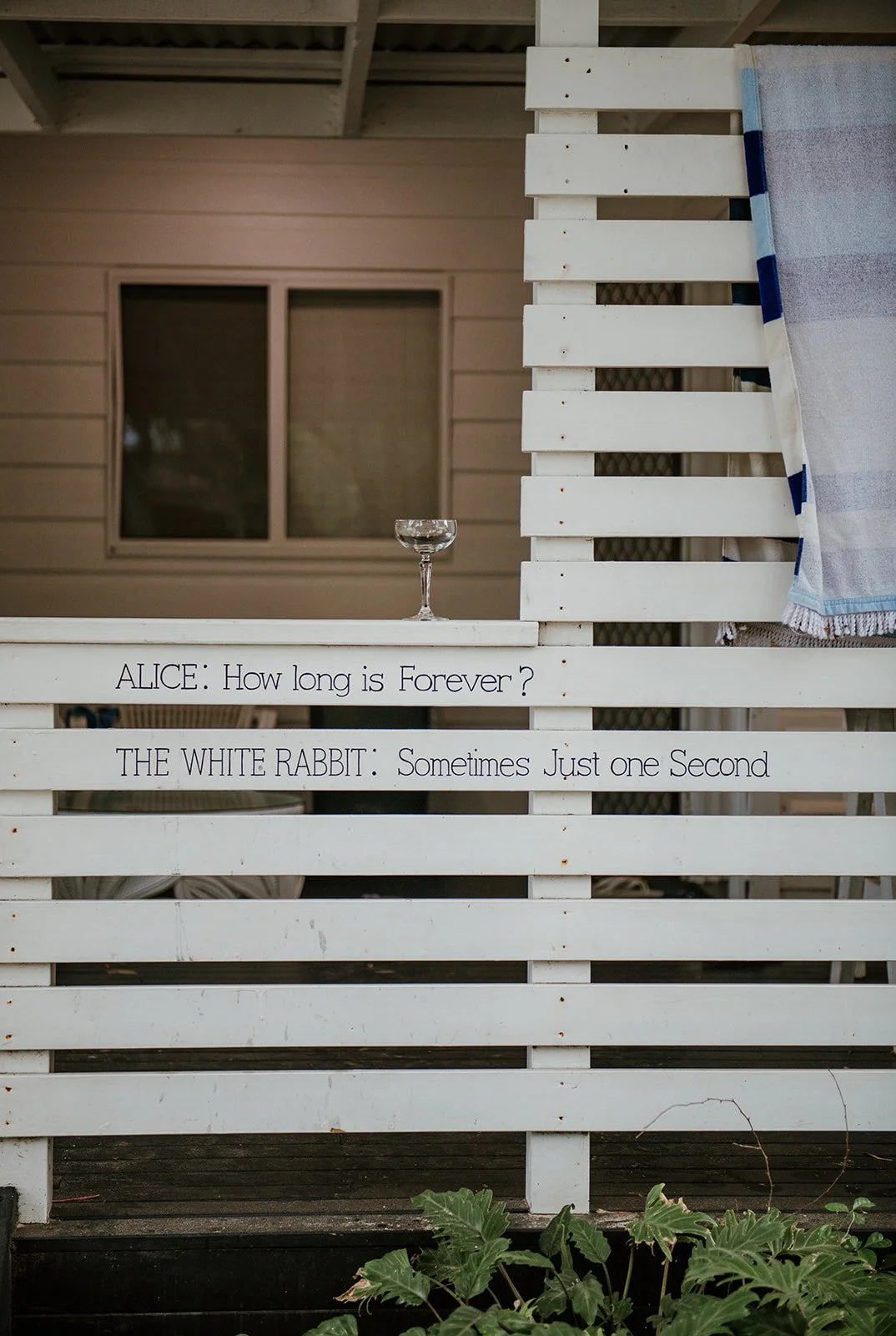 A wedding venue with a white wooden fence with a quote from Alice and the White Rabbit from Alice in Wonderland a glass on top of the fence. There are some green plants at the bottom of the fence and a house with a window in the background.
