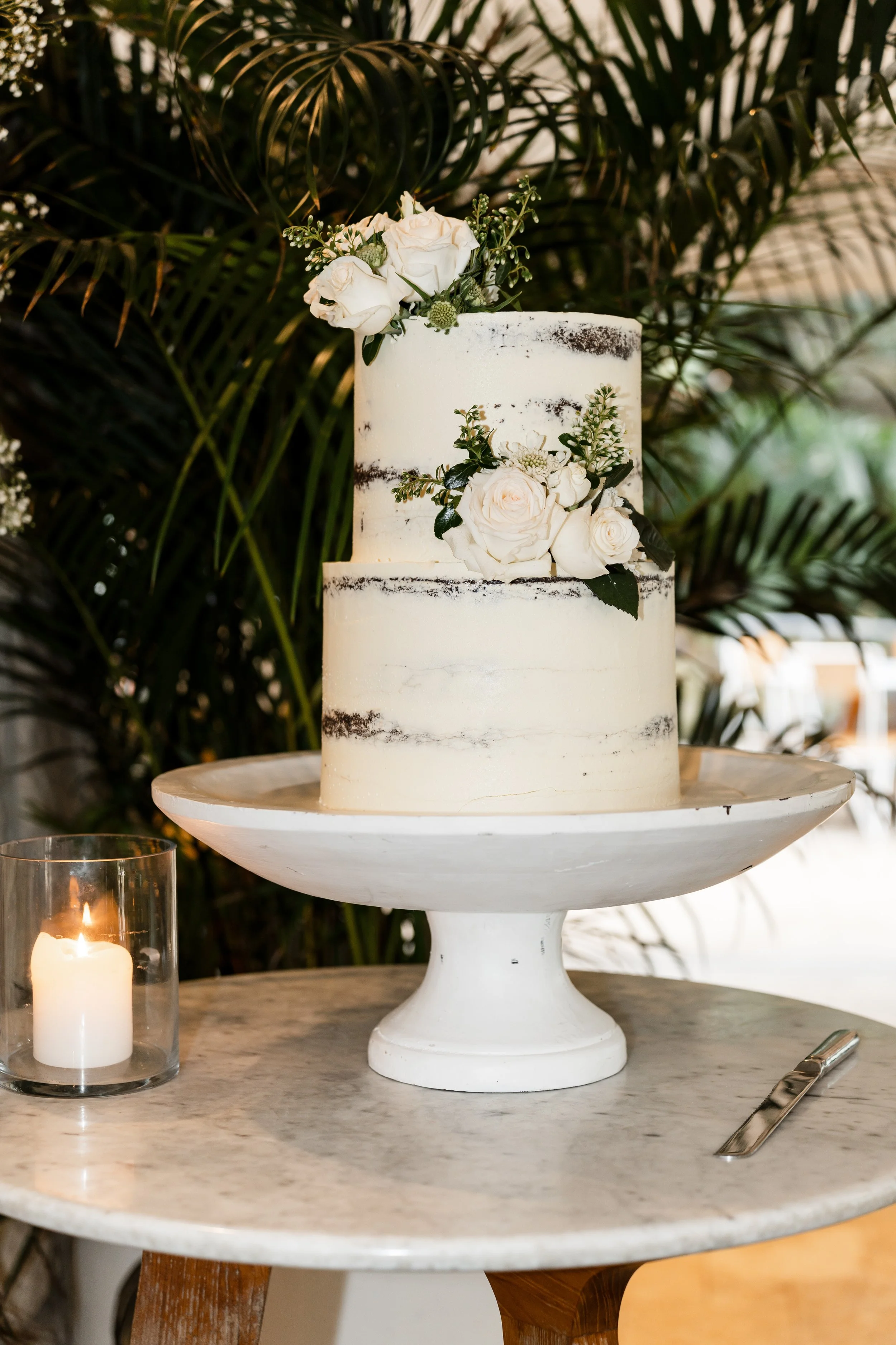 Two-tiered white wedding cake with dark chocolate streaks decorated with white roses and greenery, on a white cake stand, with a glowing candle nearby, set on a marble table in front of green leafy plants.