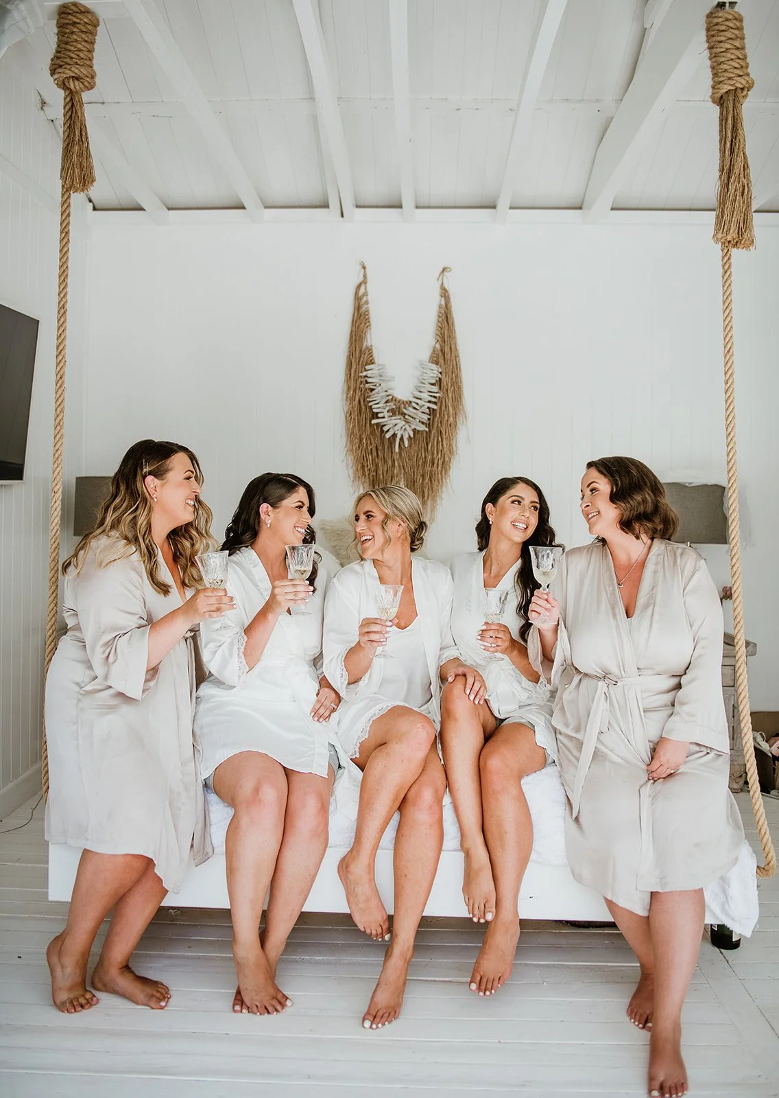 Five women dressed in white robes sitting on a bed with a swing, holding glasses, smiling, in a bright, white room with a decorative wall hanging