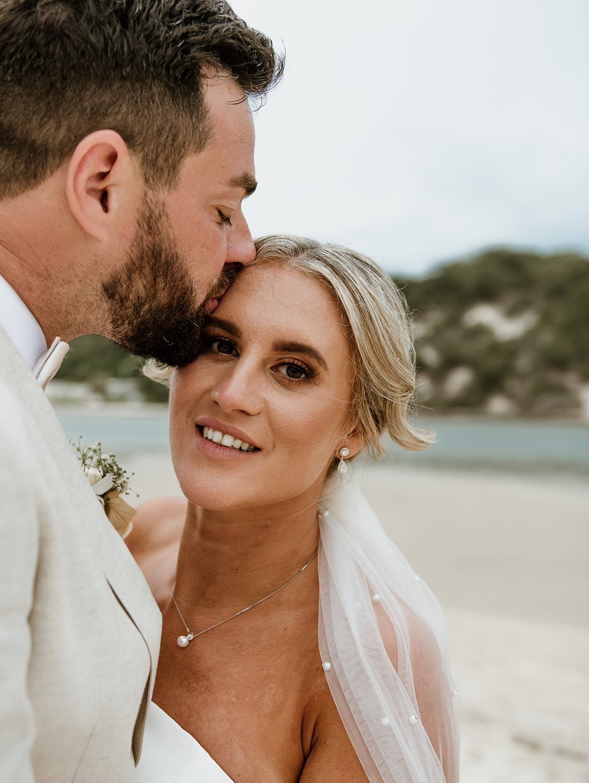 A man and woman on a beach, close-up, with the man kissing the woman's forehead. The woman has short blonde hair, is wearing earrings, a necklace, and a wedding dress with a sheer veil. The man has curly hair and a beard, and is dressed in a light-co