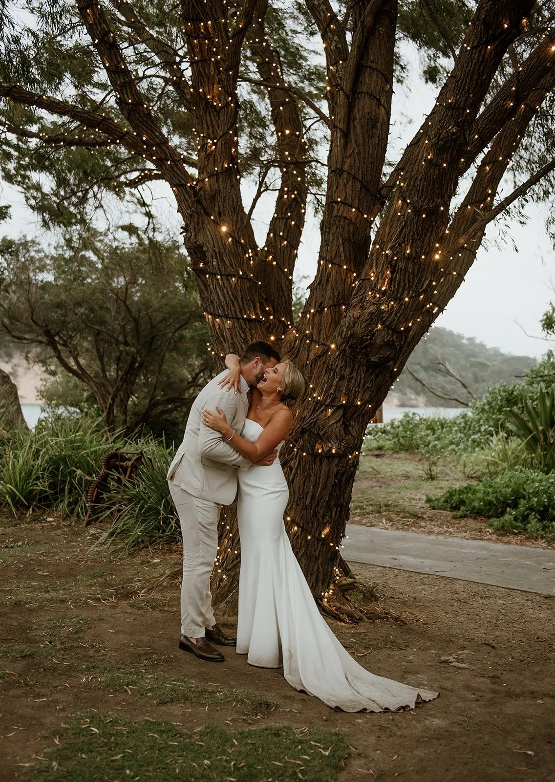 A bride and groom sharing an intimate moment by a large tree decorated with string lights, outdoors during twilight.