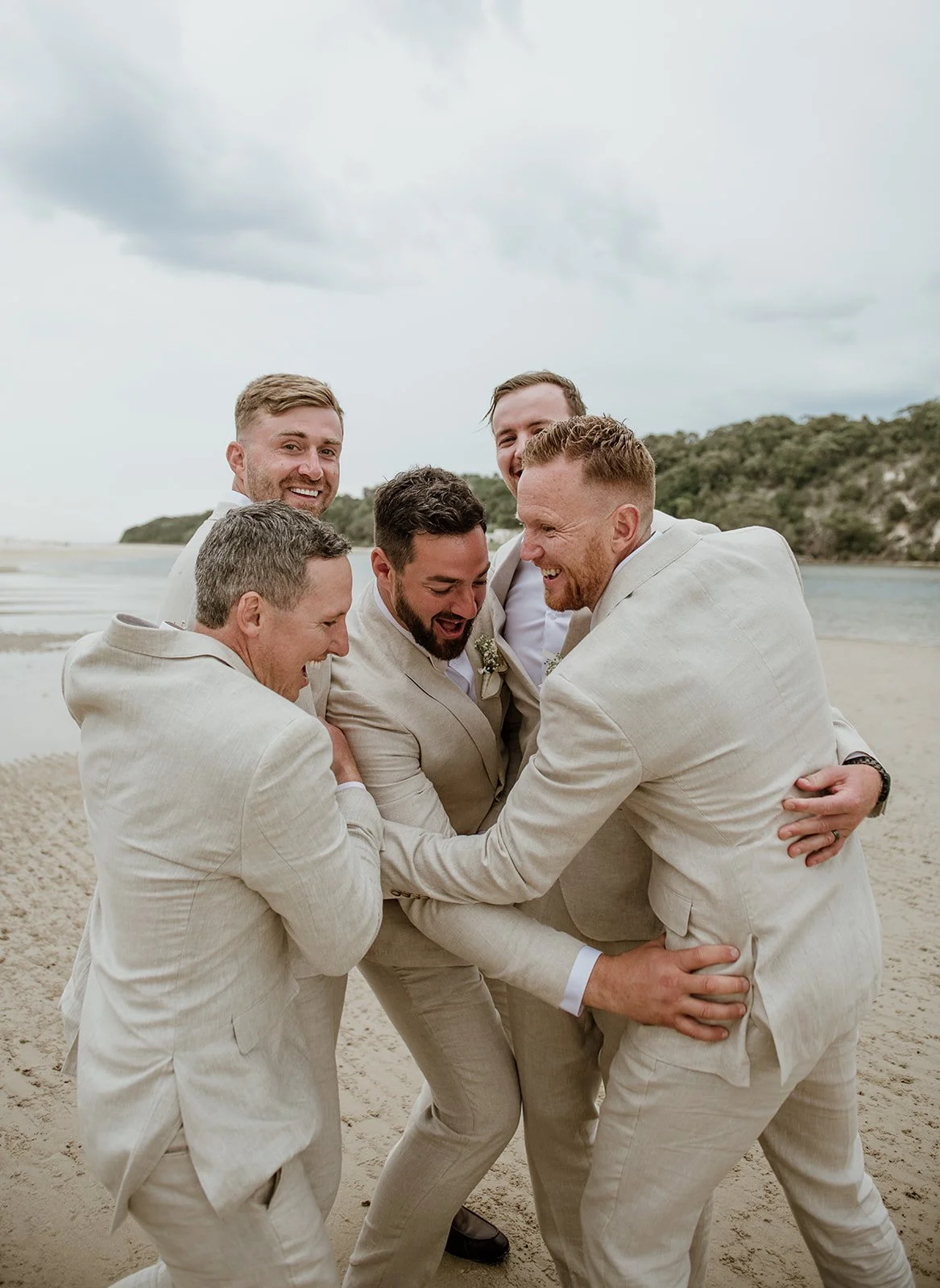Six men dressed in beige wedding suits are on a beach, tightly grouped and smiling happily.