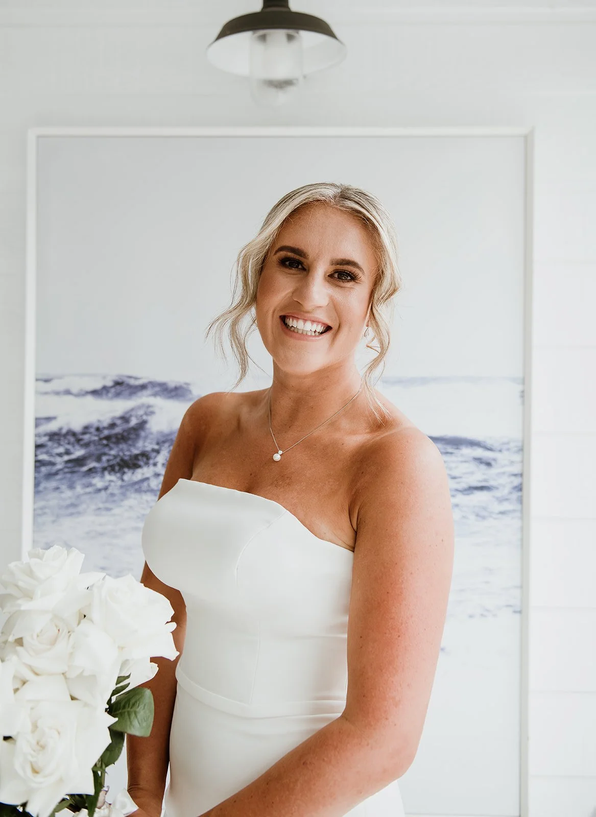 A smiling woman in a white strapless dress holding a bouquet of white roses, standing indoors with ocean art on the wall behind her.