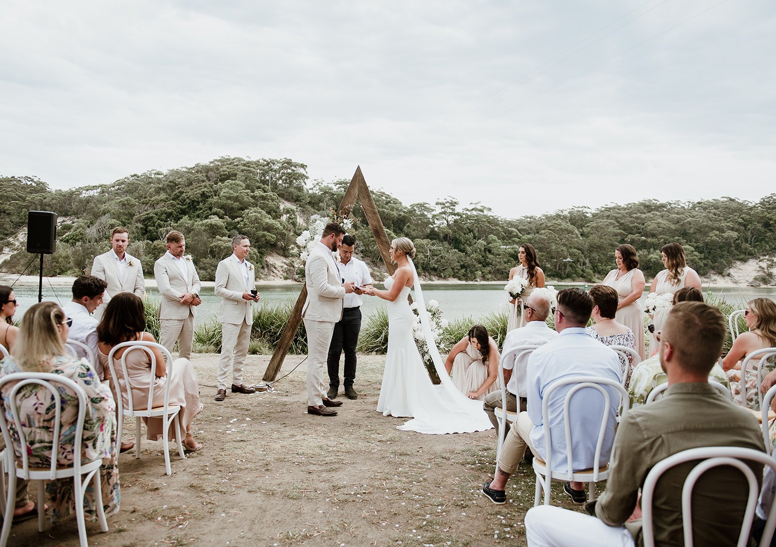 A wedding ceremony outdoors by a lake with a wooded hillside in the background, featuring a bride and groom exchanging vows under a triangular wooden arch, surrounded by bridesmaids and groomsmen, with guests seated on white chairs.
