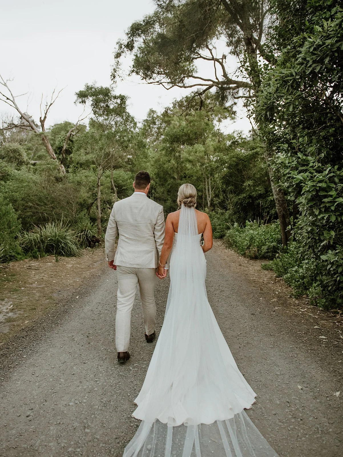 A bride and groom walking hand in hand on a dirt path surrounded by green trees and bushes, seen from behind.