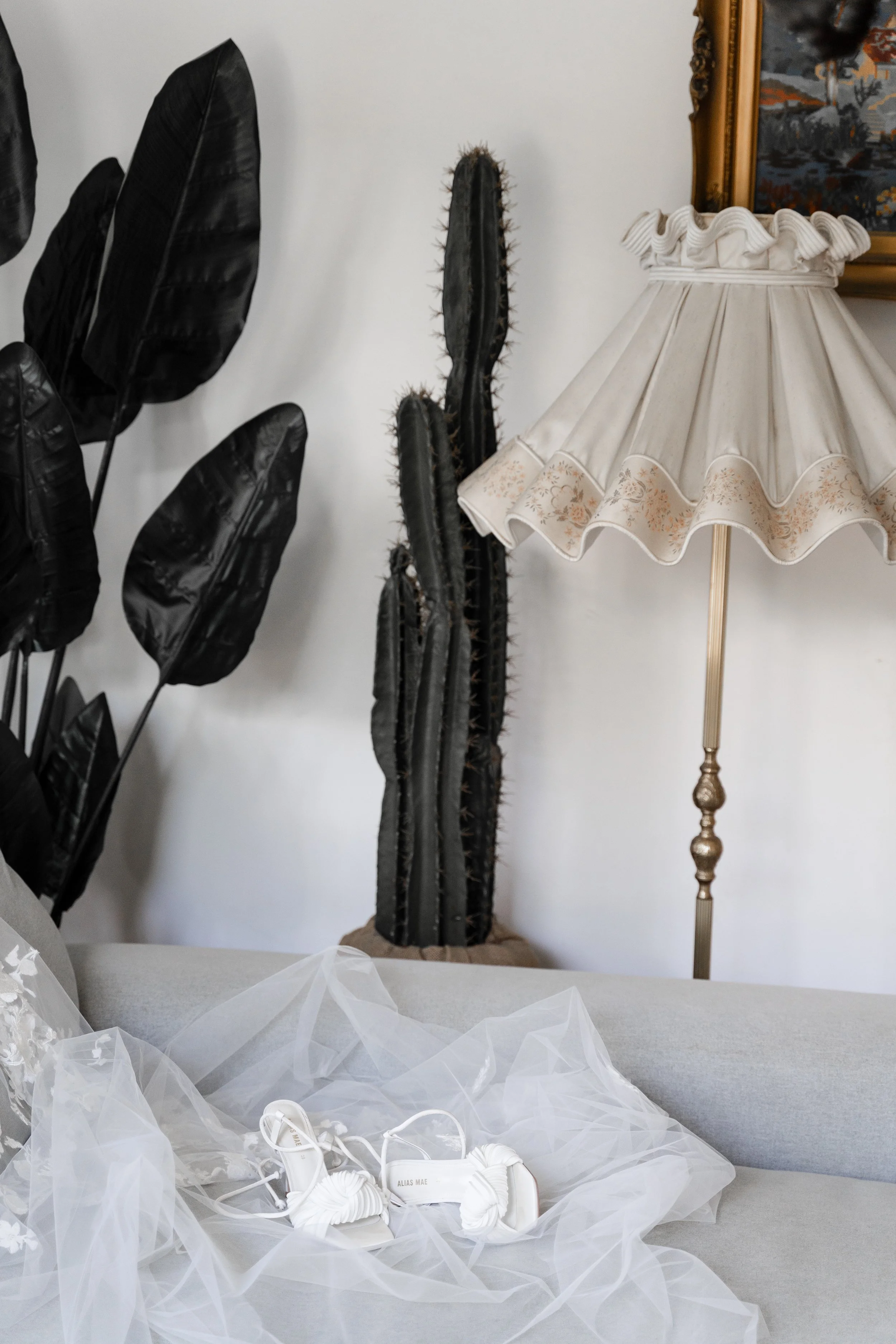 A living room corner with black wallpaper, a tall cactus, a vintage lampshade, and a wedding dress with shoes on the couch.