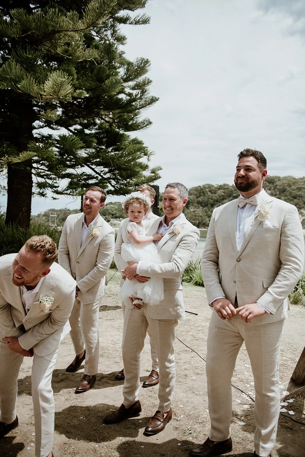 A group of men in beige suits and a young girl in a white dress at an outdoor wedding under a large tree.