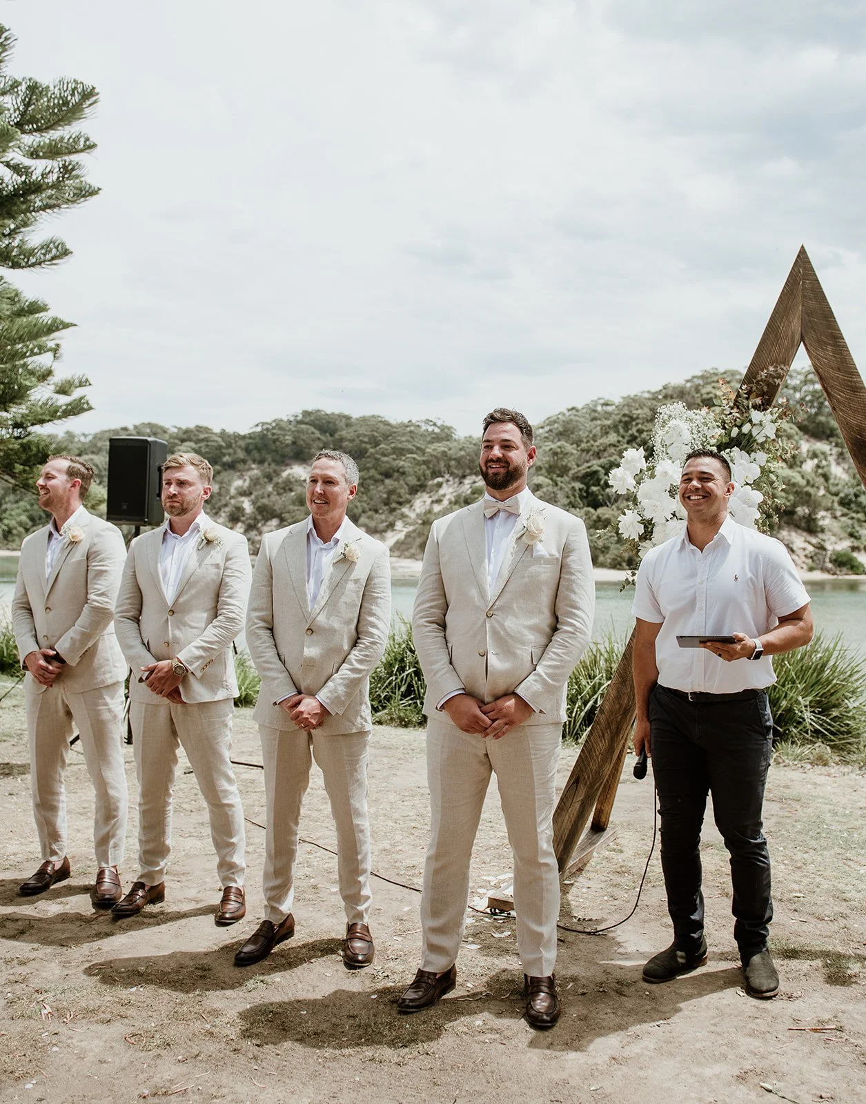 A wedding ceremony outdoors with five men standing in a row, four in beige suits and one in a white shirt with black pants, under a wooden arch decorated with white flowers, near a lake and trees.