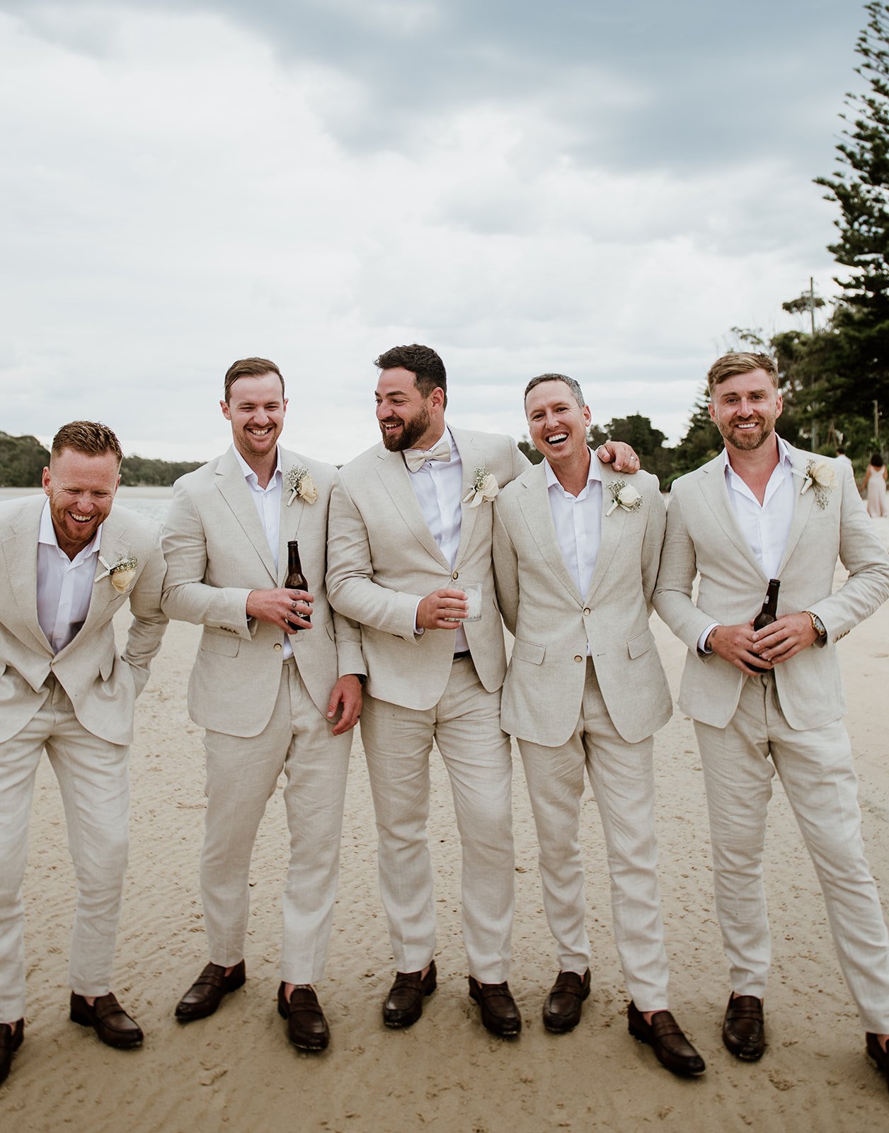 A group of five men in beige wedding suits and white shirts, smiling and laughing together on a beach, some holding drinks.