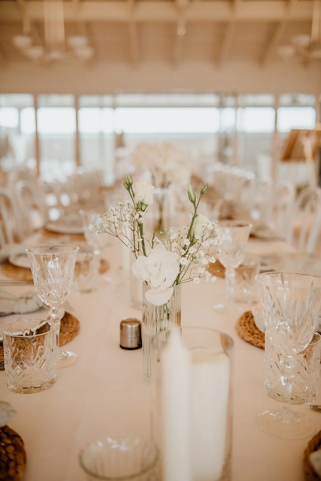 A wedding table set with glassware and a small bouquet of white flowers in a clear vase, with natural light coming in from large windows in the background.