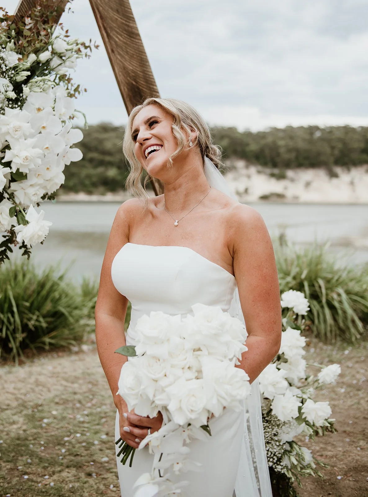 A smiling woman in a white strapless wedding dress holding a bouquet of white roses outdoors near a body of water, with a floral arch and greenery in the background.