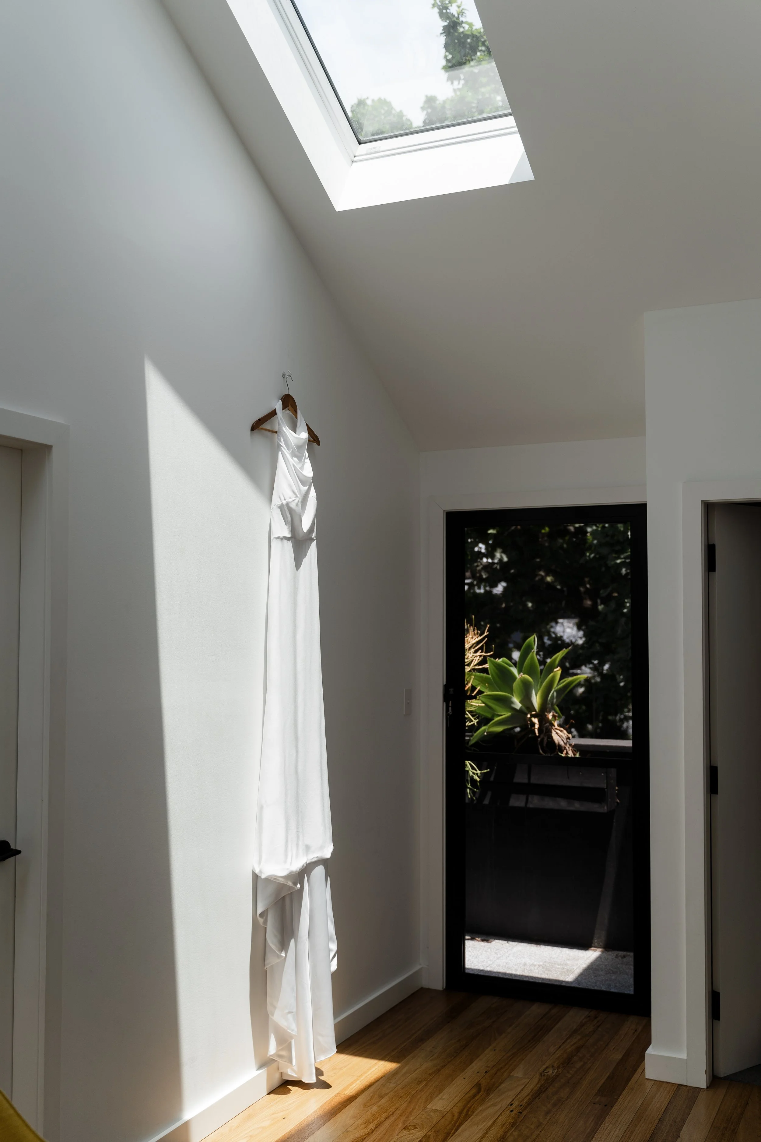 A white dress hanging on a wooden hanger on a white wall inside a bright room with a skylight and glass door leading outside, with sunlight casting shadows and a potted plant visible outside.