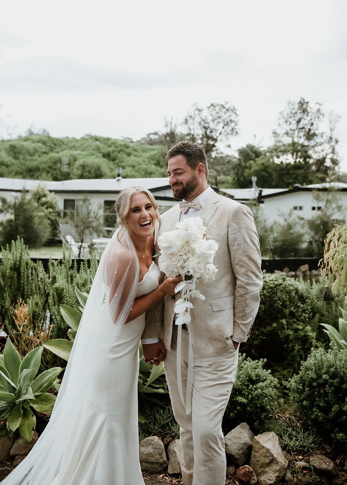 A newlywed couple smiling and holding hands outdoors, with the bride holding a cascading white bouquet, surrounded by greenery and plants.