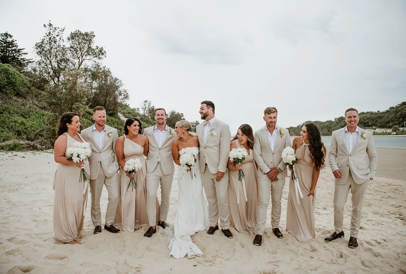 A wedding party of ten people, including the bride, groom, and bridesmaids and groomsmen, standing on a sandy beach with water and trees in the background, smiling and laughing.