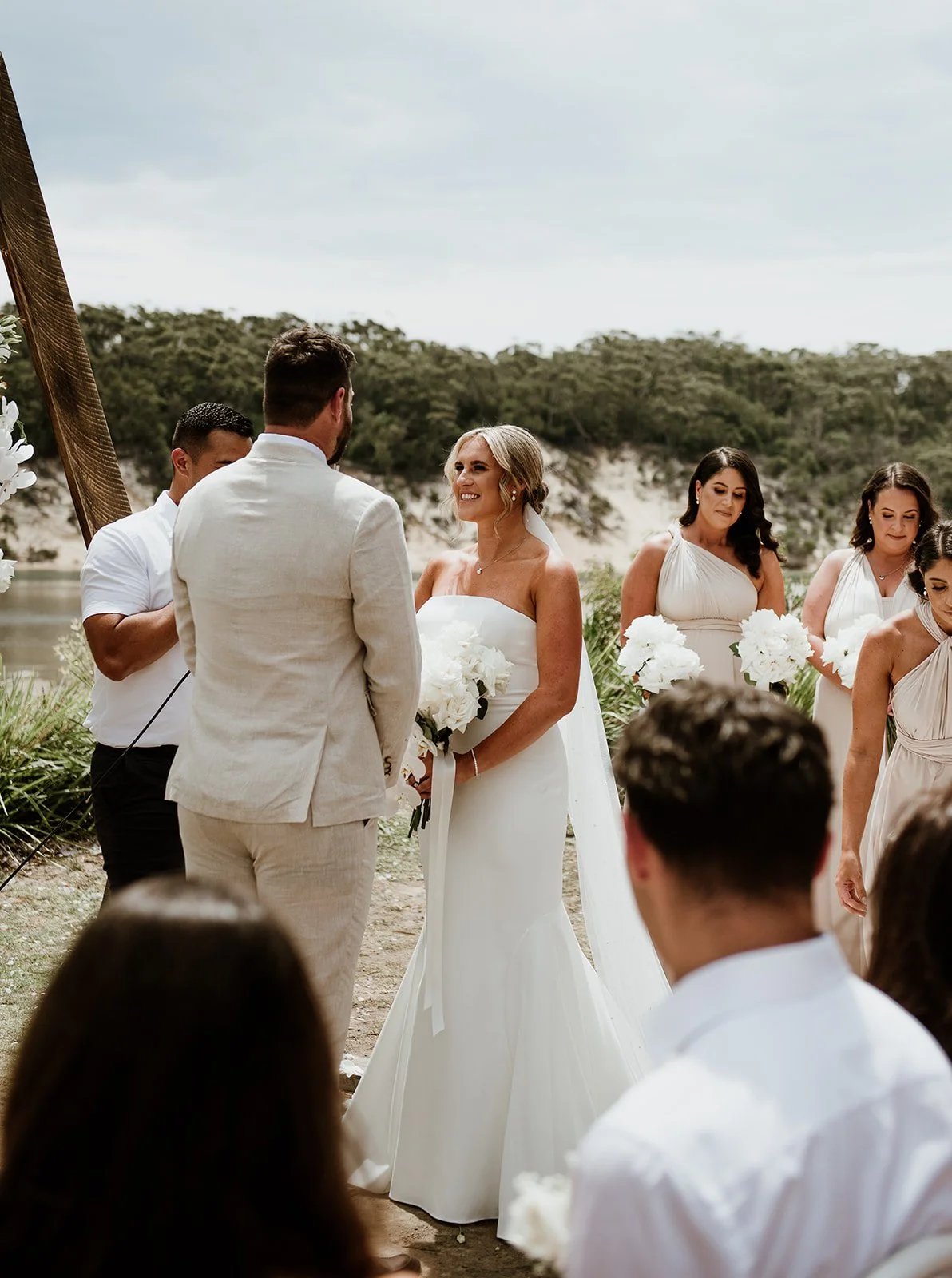 A wedding ceremony taking place outdoors by a body of water with greenery in the background. The bride, in a white strapless dress, stands holding a bouquet, facing the groom, who is dressed in a light-colored suit. Bridesmaids in matching dresses an
