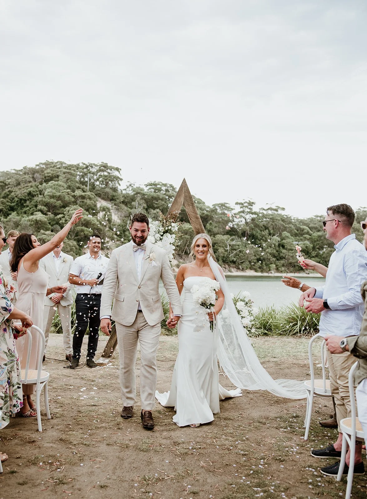 A bride and groom walking hand in hand at their outdoor wedding ceremony by a lake, surrounded by friends and family, with a triangular wooden arch behind them.