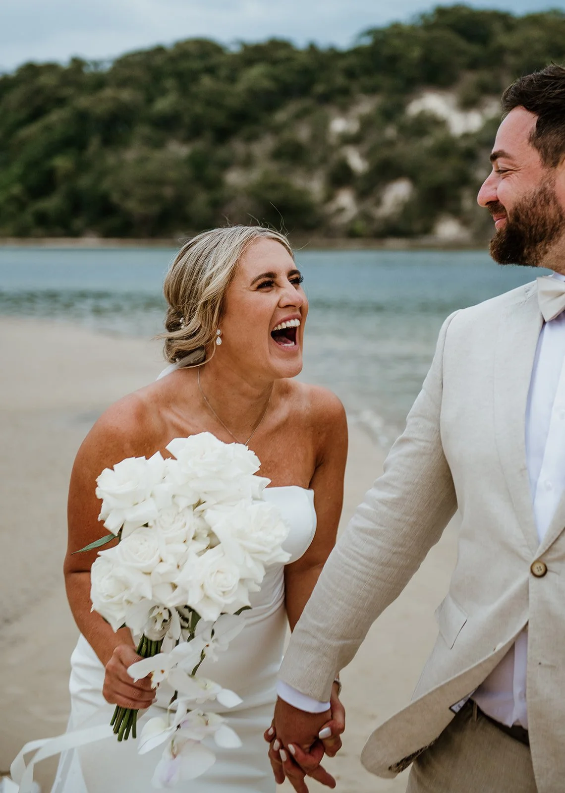 A woman in a white wedding dress holding a white bouquet, laughing while holding hands with a man in a light beige suit, on a beach with water and green hills in the background.