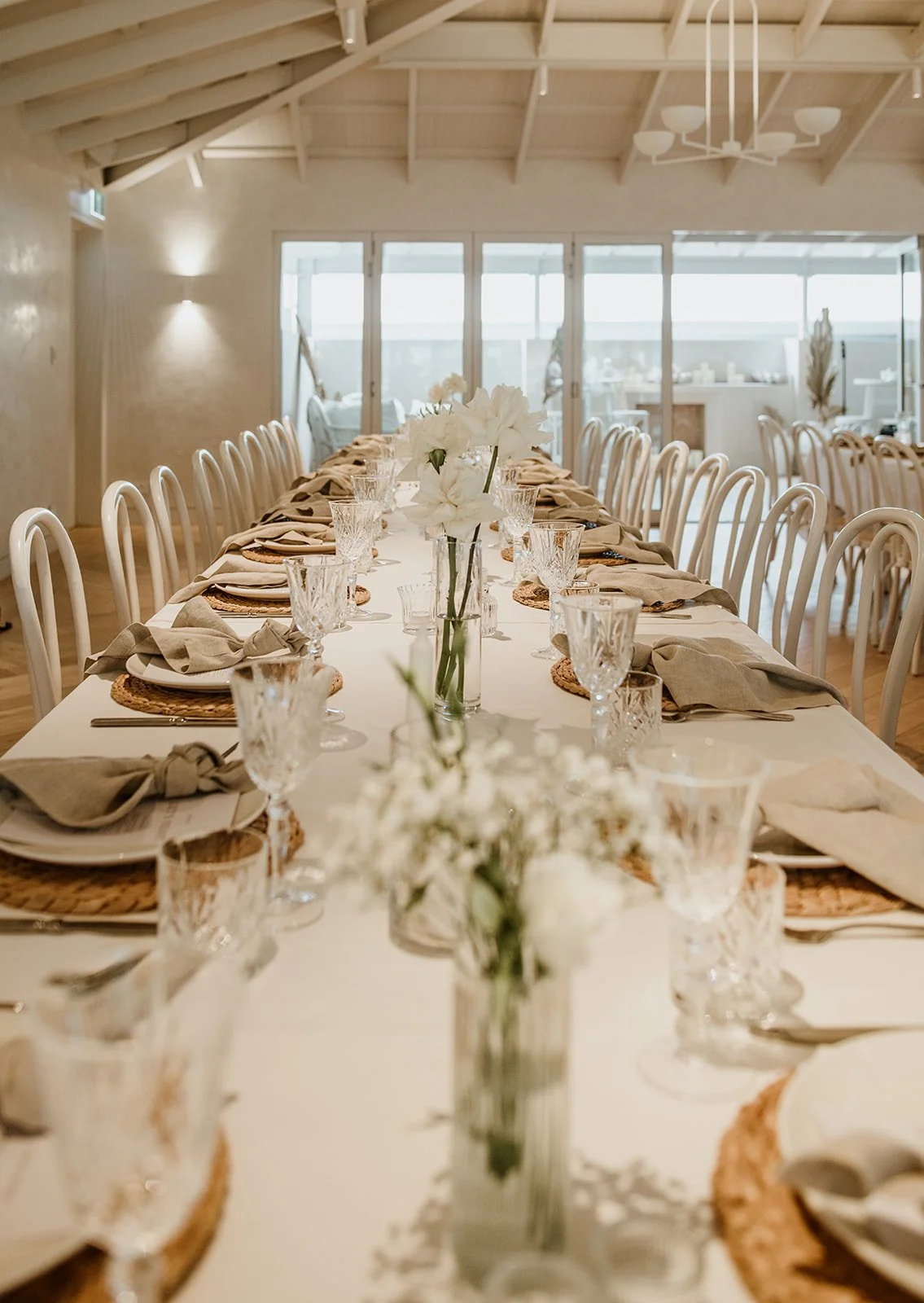 A long wedding dining table decorated with white flowers in vases, crystal glasses, beige napkins, and woven placemats in a bright, airy room with beige walls and large windows.
