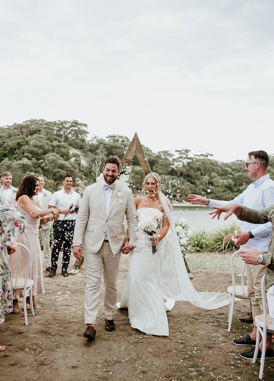A bride and groom walking hand in hand at their outdoor wedding ceremony, surrounded by friends and family, with trees and a lake in the background, as confetti is thrown in celebration.