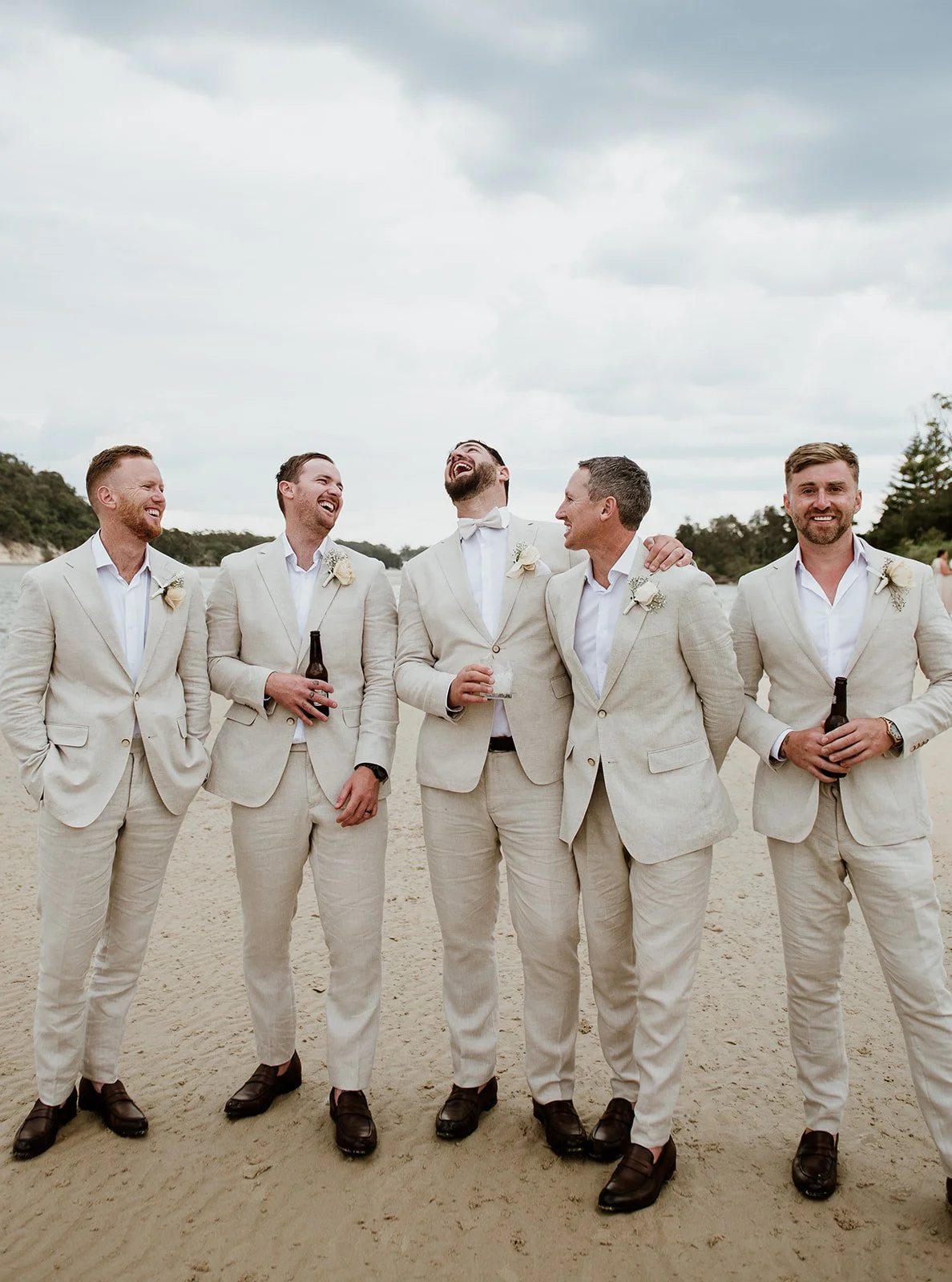 Six men in beige suits standing on a sandy beach, laughing and holding drinks, during a wedding celebration.