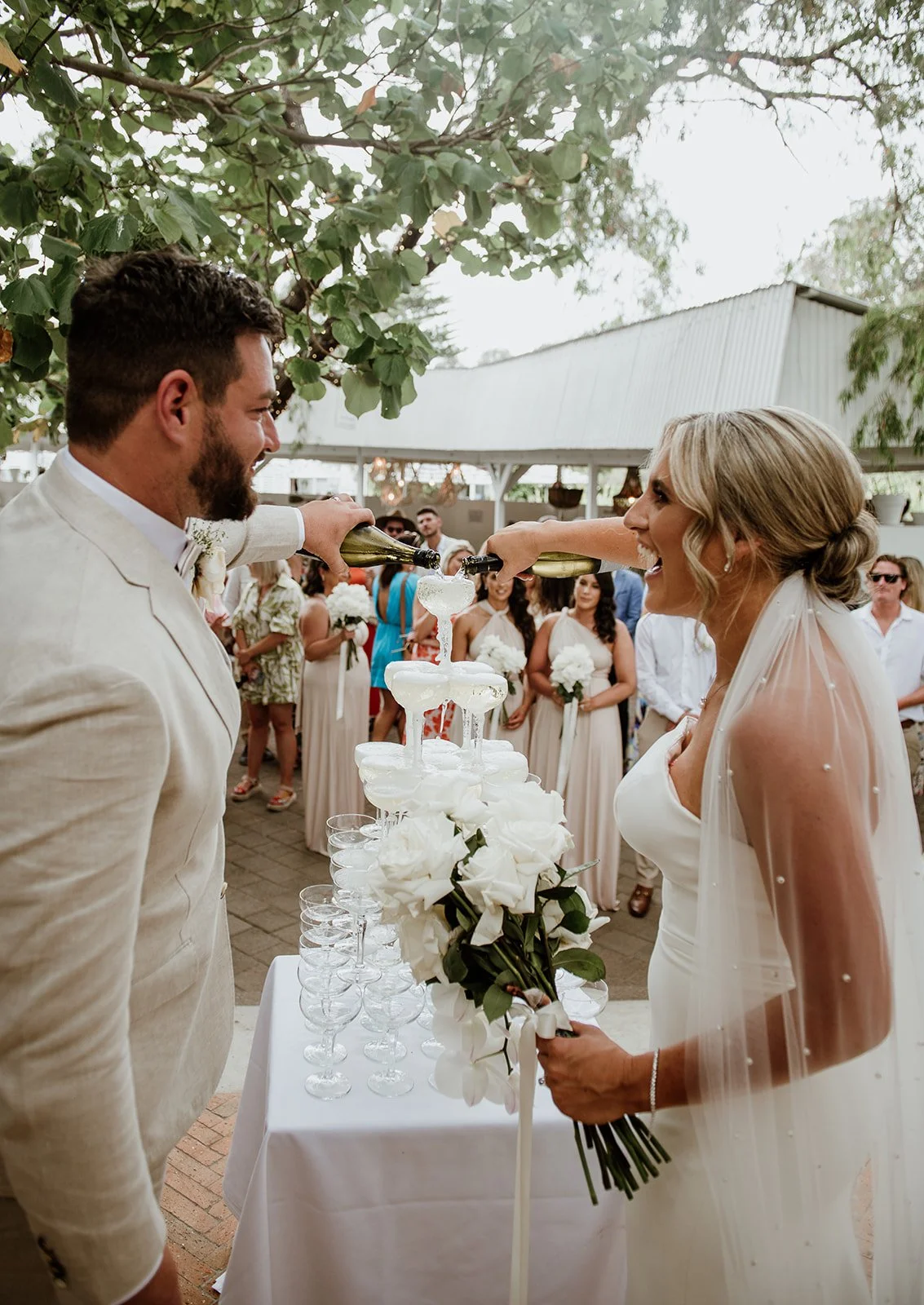 A bride and groom are pouring champagne over a champagne tower at their wedding reception, surrounded by wedding guests.