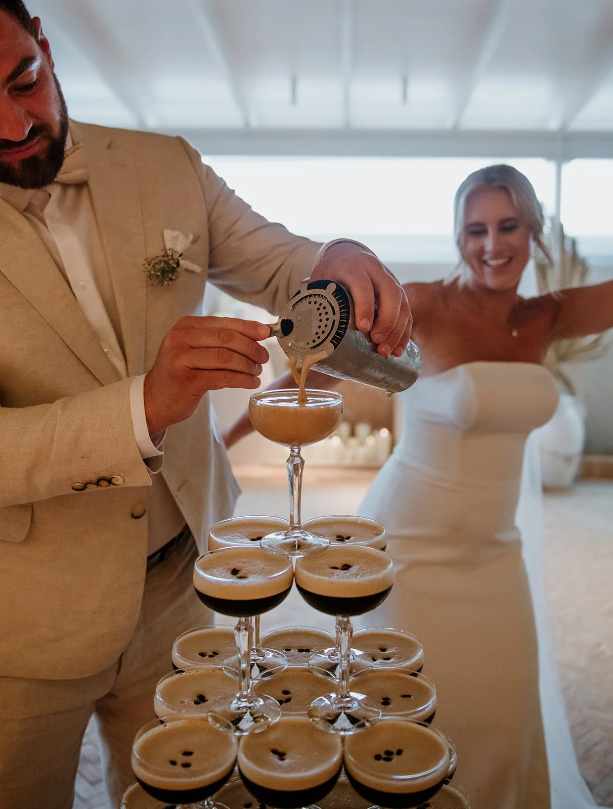 A bride and groom celebrating with a tower of cocktails, the groom pouring a drink into a glass, both dressed in wedding attire, smiling in a bright, elegant setting.