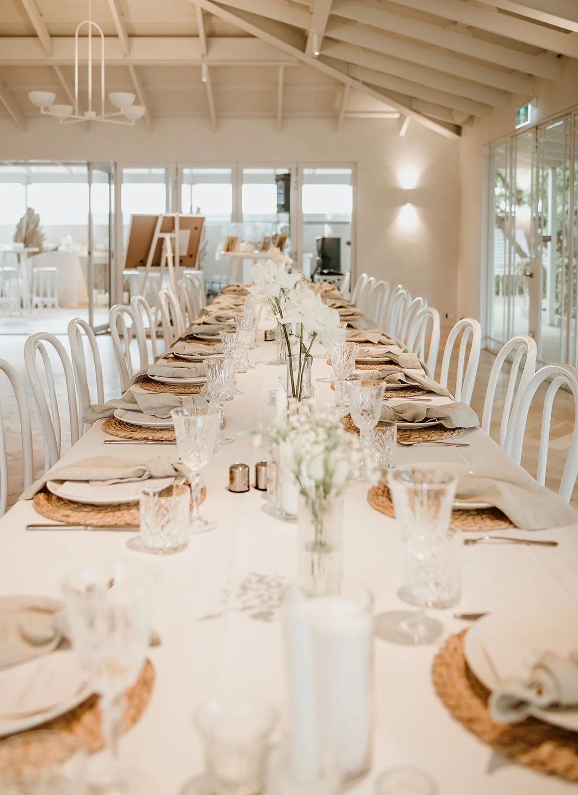 Long dining table set for a formal event with white floral centerpieces, glasses, napkins, and woven placemats in a bright, airy room with large windows and a high, peaked ceiling.