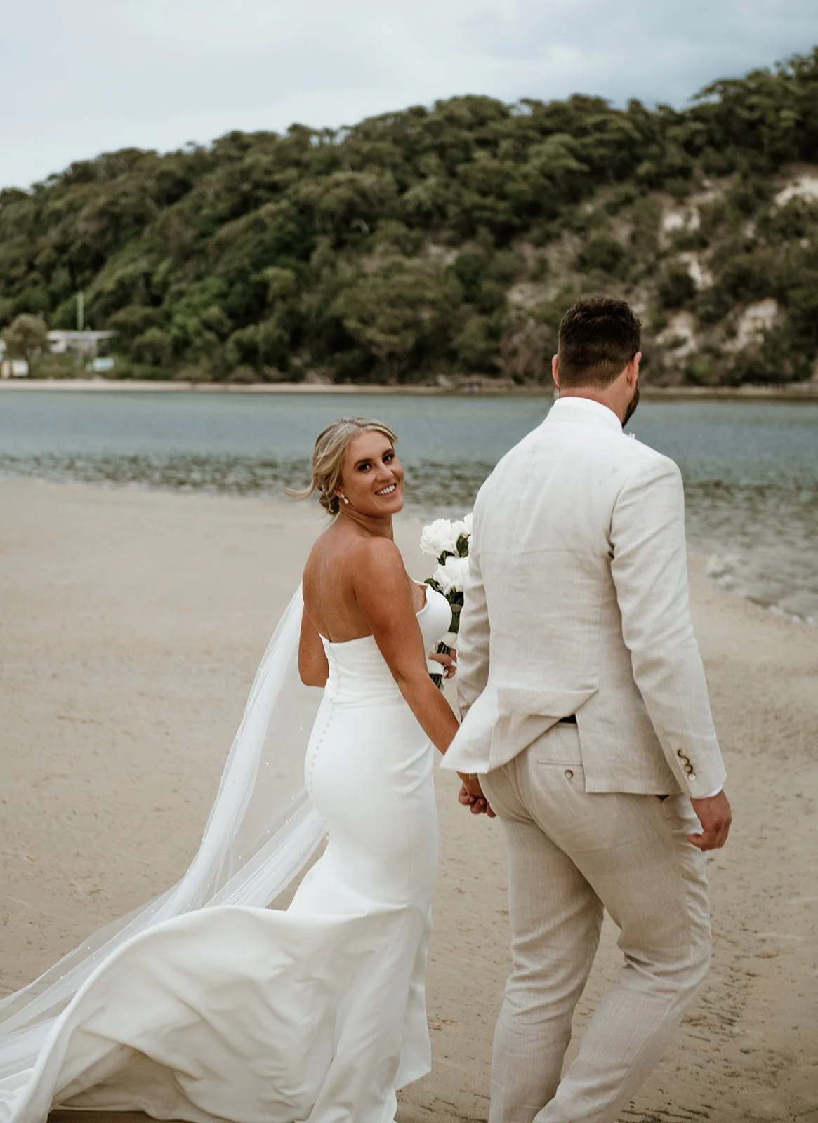 A bride and groom holding hands on a beach, with the bride smiling and looking back at the camera, beach and water in the background.