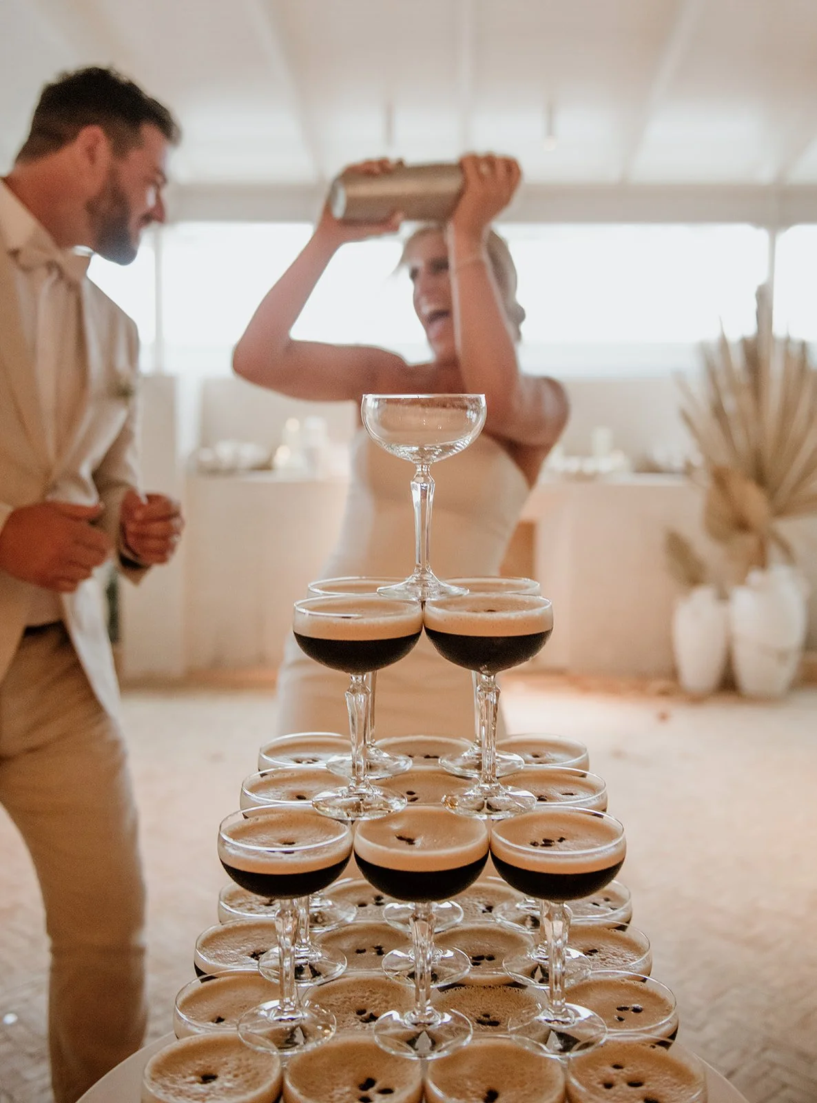 A table with a pyramid of glasses filled with black coffee or espresso, with two people dancing and celebrating in the background.