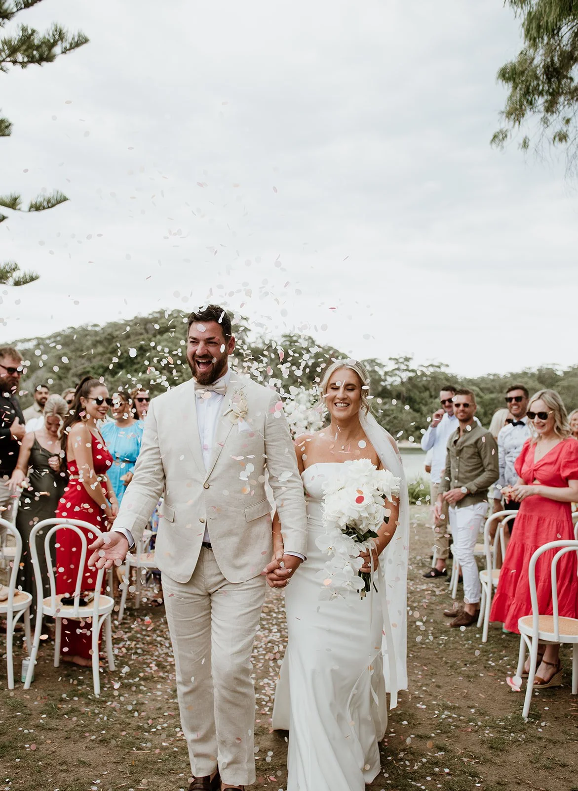 A newlywed couple walks hand-in-hand through confetti at an outdoor wedding ceremony, surrounded by smiling guests.
