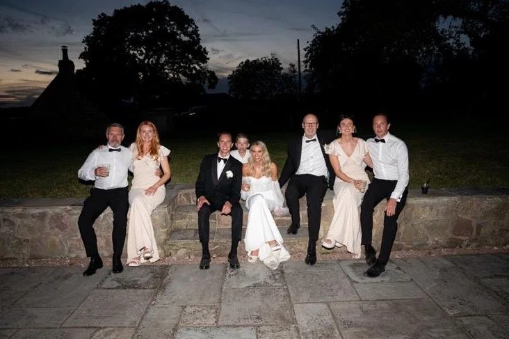 Group of seven people dressed in formal attire, sitting on a stone wall outdoors during dusk, with trees and a building in the background.