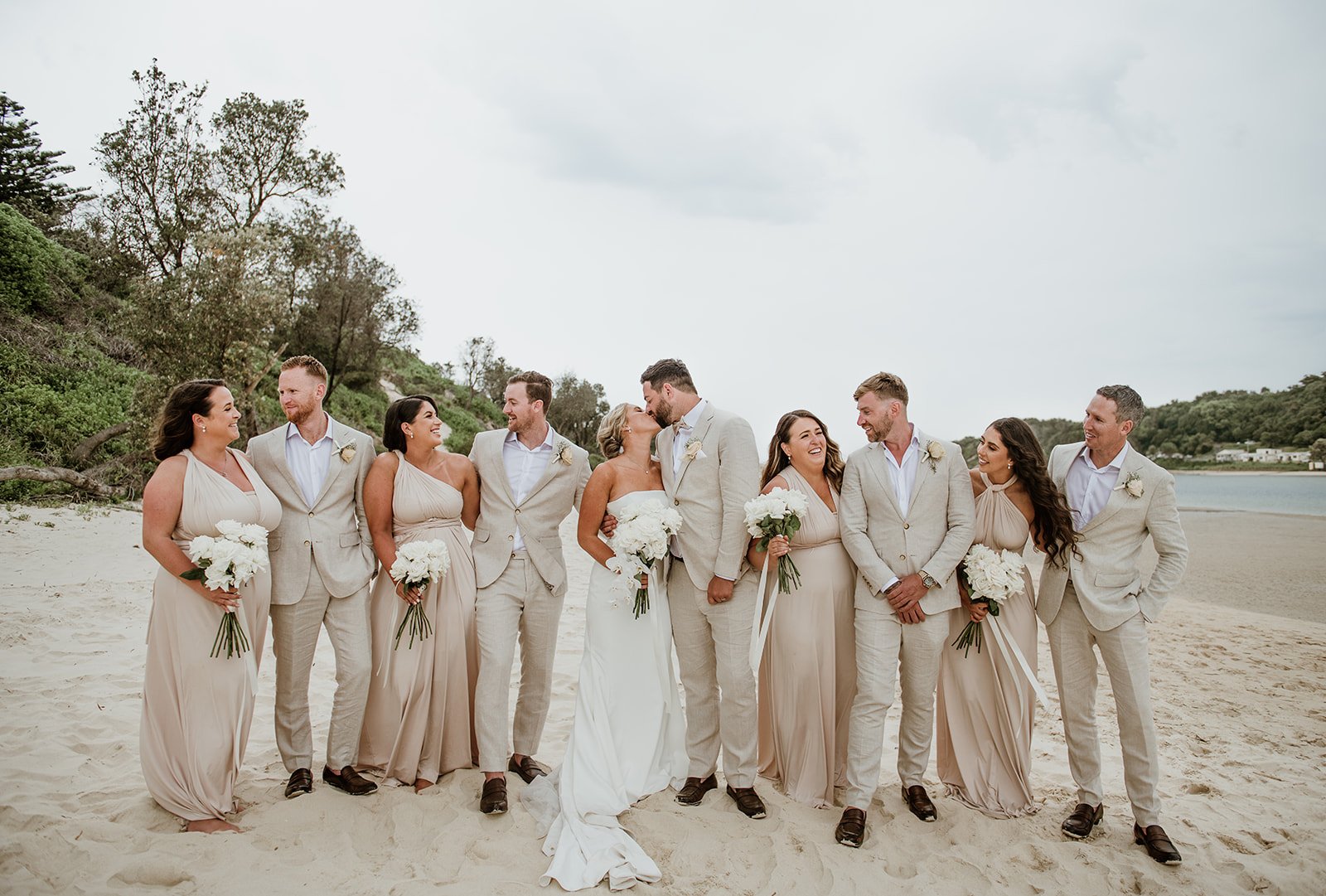 A wedding party of ten people, including the newlywed couple, standing on a sandy beach holding white bouquets, with trees, water, and a cloudy sky in the background.