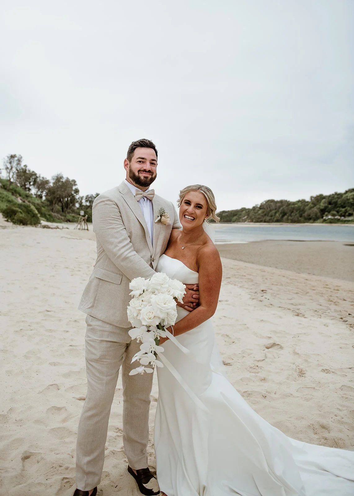 Bride and groom standing on a beach, smiling, with the bride holding a bouquet of white flowers.