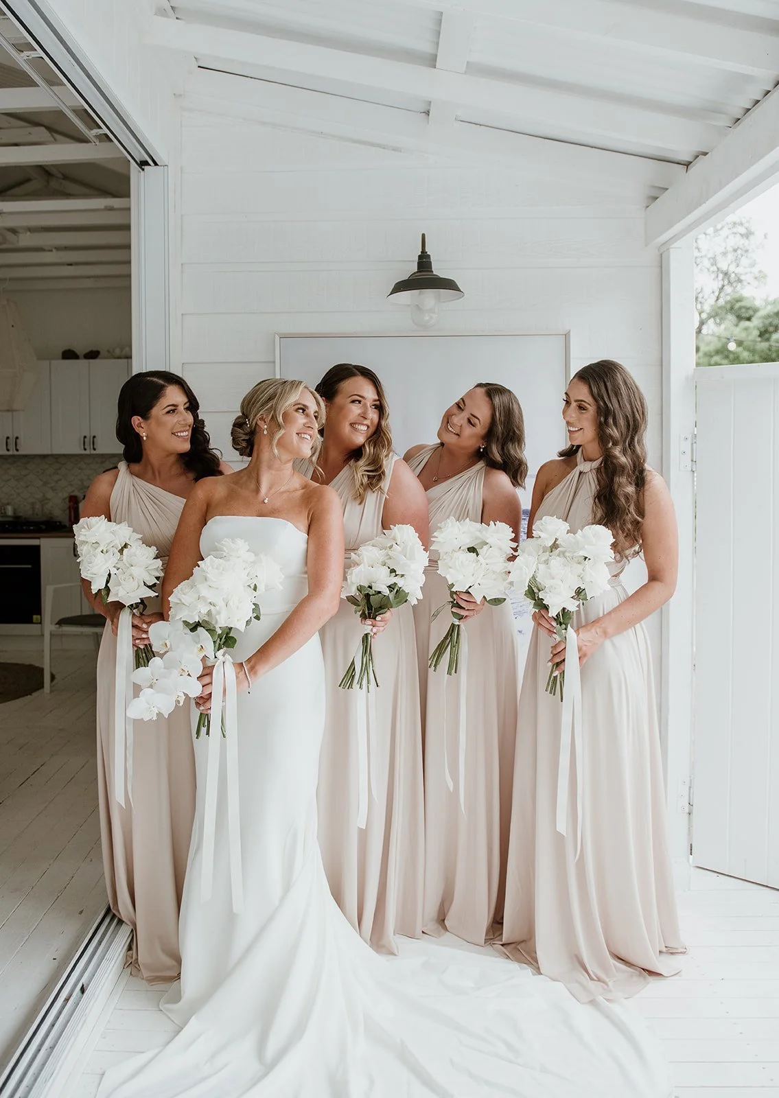 Bride and four bridesmaids in a bright, white room with white walls, holding white floral bouquets, smiling and looking at each other, all wearing cream-colored dresses for a wedding.