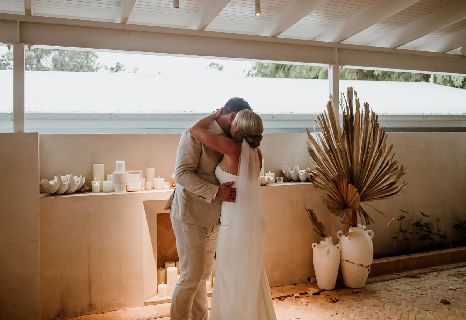A couple sharing a kiss at their wedding, with candles and large vases with dried palm leaves in the background.