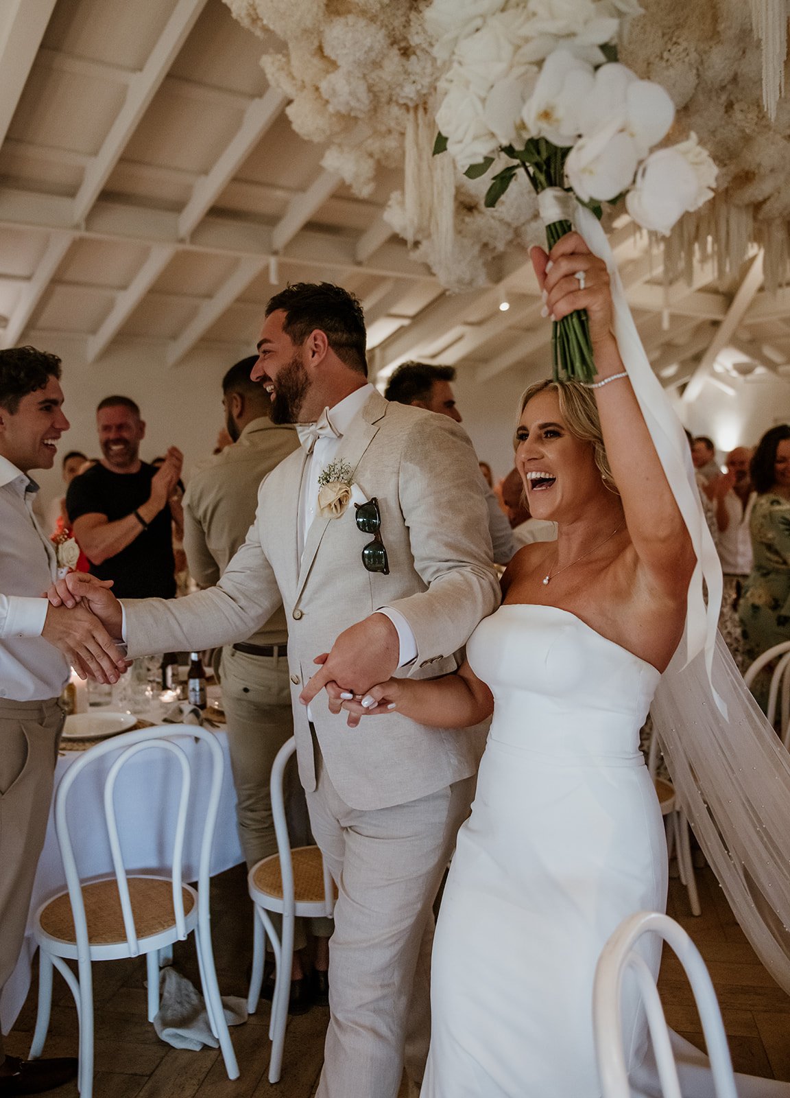 A bride holding a bouquet of white flowers and wearing a strapless white wedding dress, standing next to a groom in a beige suit, at a wedding reception.
