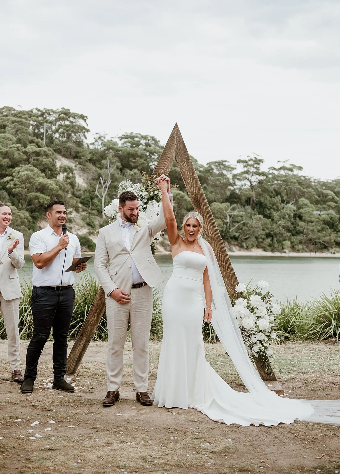 A newly married couple celebrates with their arms raised during an outdoor wedding ceremony by a river, with green trees and cloudy sky in the background.