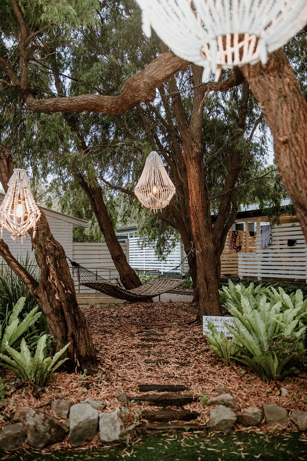 A backyard scene with a large tree decorated with hanging lights, a hammock tied between two trees, and a pathway lined with plants and stones leading to the hammock. There are outdoor structures like a white fence and a small shed in the background.