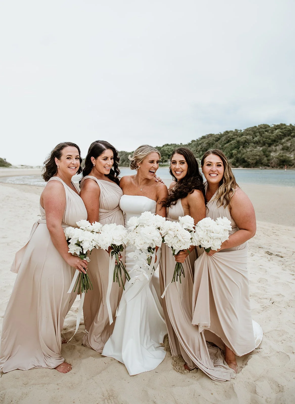 A bride and five bridesmaids standing on a sandy beach near water with a hilly landscape in the background. The women are smiling and holding white bouquets with large white flowers, dressed in beige or champagne-colored dresses.