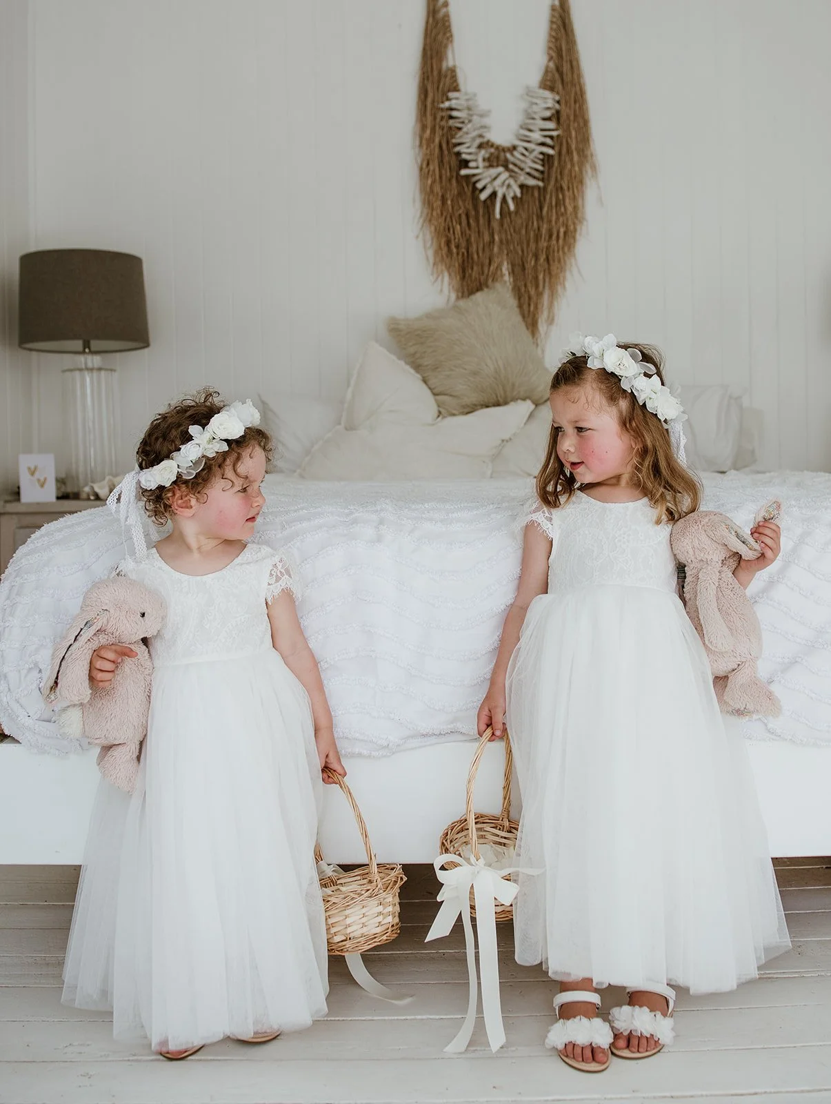 Two young girls dressed in white lace dresses and flower crowns, holding stuffed bunnies and small baskets, standing in a cozy bedroom with a bed, pillows, and wall hanging in the background.